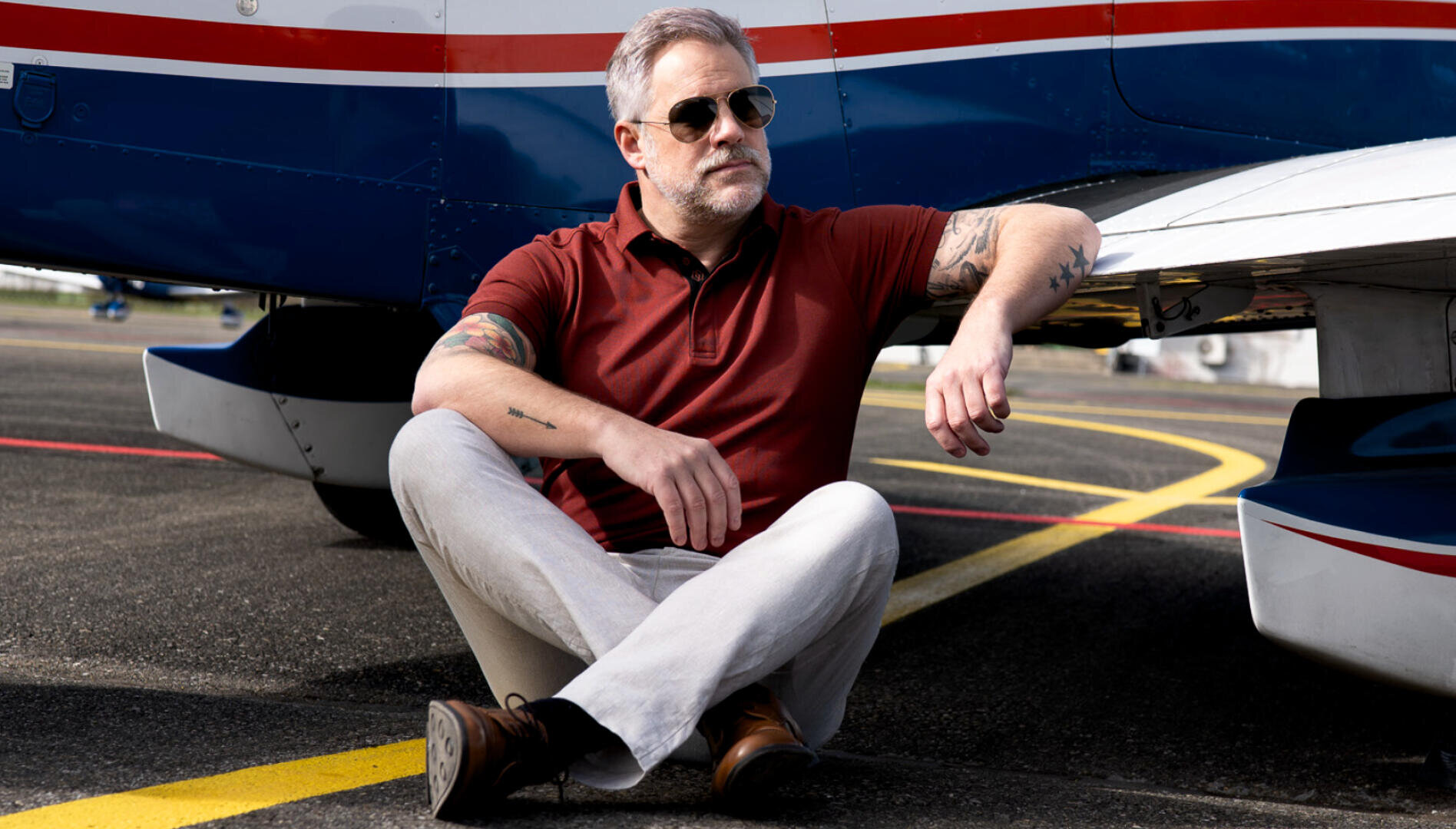 Ein Mann mit grauem Haar und Bart, der eine Sonnenbrille, ein rotes Polohemd und eine helle Hose trägt, sitzt im Schneidersitz auf der Rollbahn neben einem blau-weißen Flugzeug.