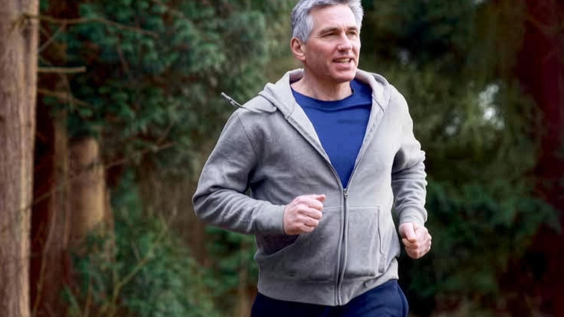 Ein Mann mittleren Alters mit grauen Haaren joggt im Freien in einem Waldgebiet, er trägt einen grauen Kapuzenpullover mit Reißverschluss und ein marineblaues Hemd, im Hintergrund sind Bäume und Grünflächen zu sehen.