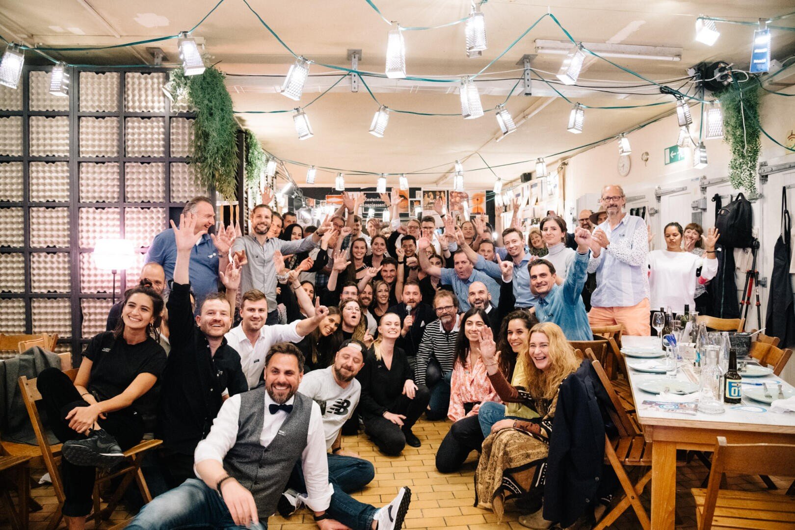 Eine große Gruppe von Menschen posiert und lächelt für ein Foto in einem hell erleuchteten Restaurant. Sie sitzen und stehen dicht beieinander zwischen Tischen und Holzstühlen, über denen Lichterketten und Grünpflanzen hängen.