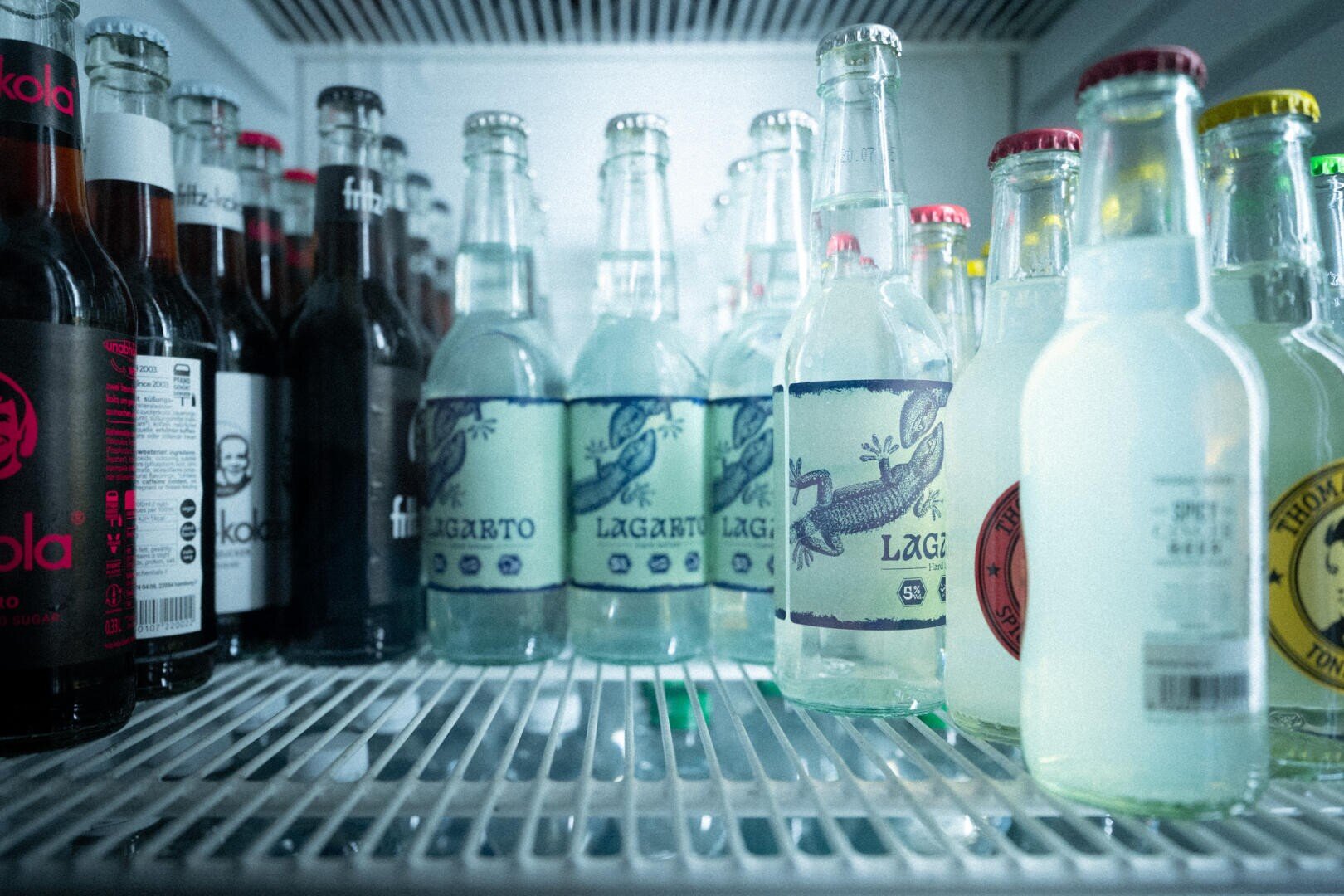 Verschiedene Glasflaschen mit Getränken, darunter Limonaden und kohlensäurehaltige Getränke, sind aufrecht auf einem Kühlregal angeordnet. Etiketten mit bunten Motiven und Logos sind zu sehen, und Kondenswasser zeigt an, dass die Getränke kalt sind.