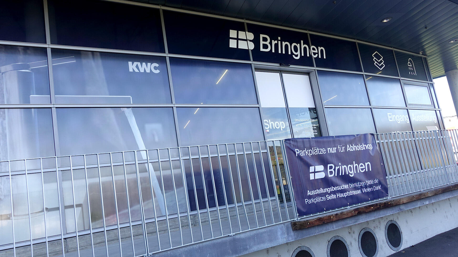 Ein Schaufenster mit großen Fenstern und Schildern mit der Aufschrift Bringhen, KWC und Shop. Ein Banner davor weist darauf hin, dass der Parkplatz nur für Bringhen-Kunden bestimmt ist. Ein Metallgeländer verläuft entlang der Eingangsplattform.