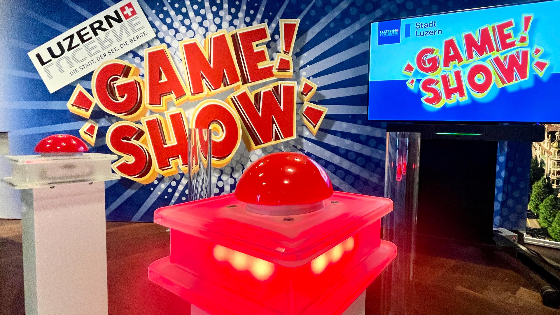 Ein grosser roter Game-Show-Buzzer auf einem beleuchteten Podium steht vor einem farbenfrohen GAME SHOW-Schild und einem Bildschirm mit der Aufschrift Stadt Luzern GAME SHOW in einer hellen, energiegeladenen Studioumgebung.