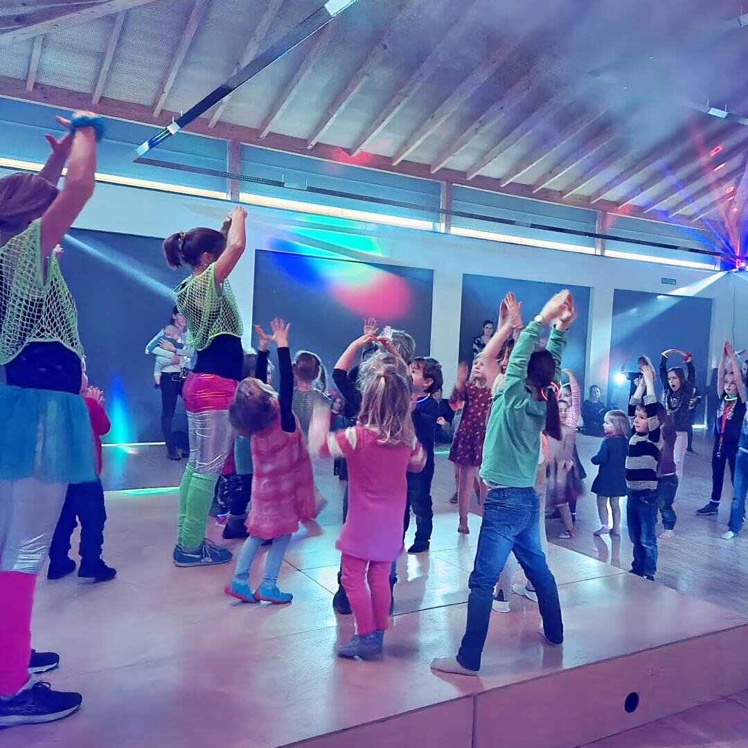 Kinder und Erwachsene tanzen energisch auf einer Bühne unter bunten Lichtern in einem geräumigen Raum mit Holzdecke und schaffen eine lebendige und fröhliche Atmosphäre.