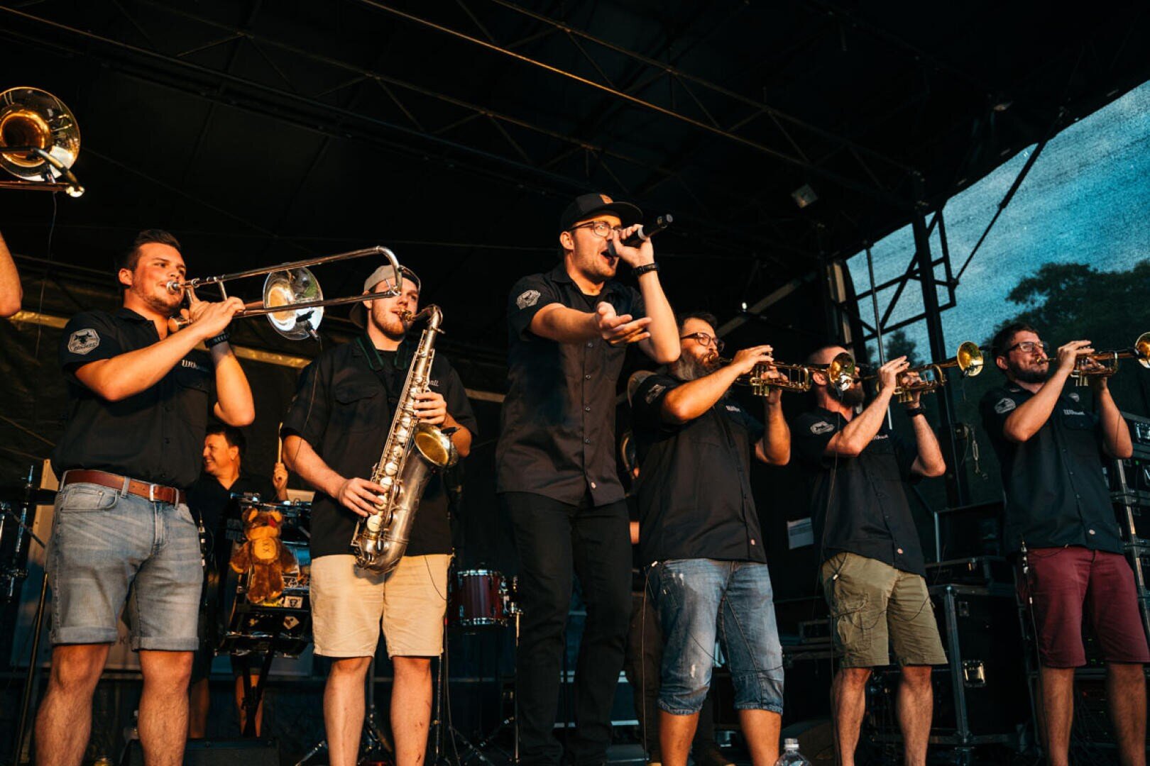 Auf der Bühne tritt eine lebhafte Band auf, bestehend aus Musikern, die Blasinstrumente und ein Saxophon spielen, sowie einem Sänger mit Mikrofon. Sie tragen passende dunkle Hemden und Shorts und schaffen eine energiegeladene Atmosphäre bei einer Veranstaltung im Freien.
