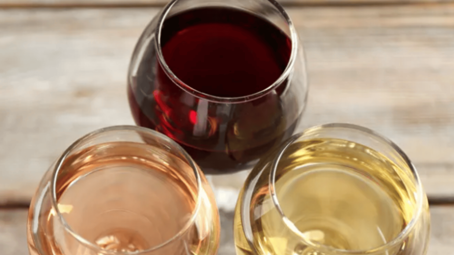 Drei Weingläser - ein rotes, ein weißes und ein roséfarbenes - sind in einem Dreieck auf einer rustikalen Holzfläche angeordnet, von oben betrachtet.