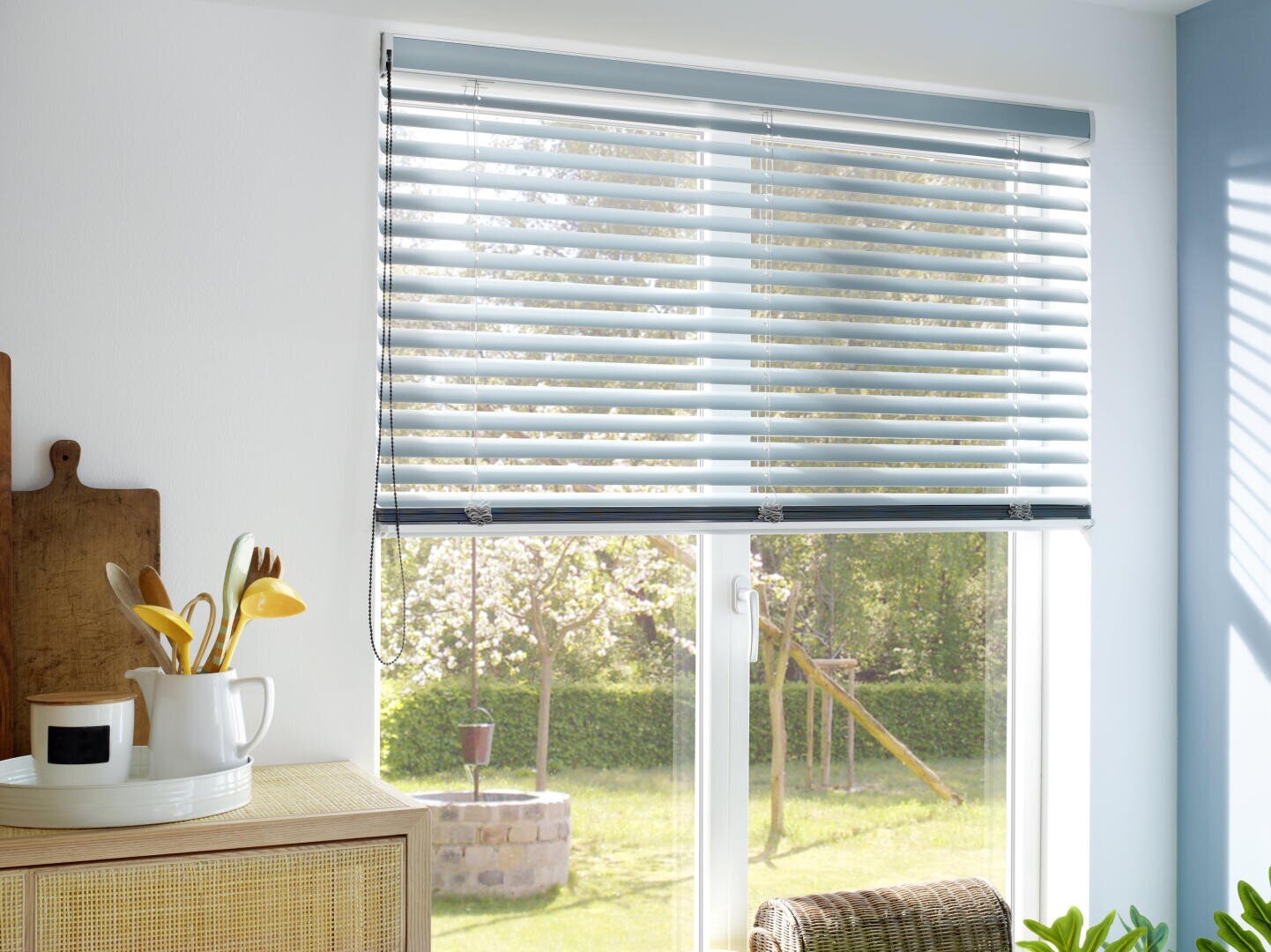 Eine sonnenbeschienene Küche mit einem Fenster, das von hellblauen horizontalen Jalousien bedeckt ist und einen grünen Garten mit einer Schaukel zeigt. Im Inneren befindet sich ein Holzschrank mit Küchenutensilien und ein Korbstuhl in der Nähe.