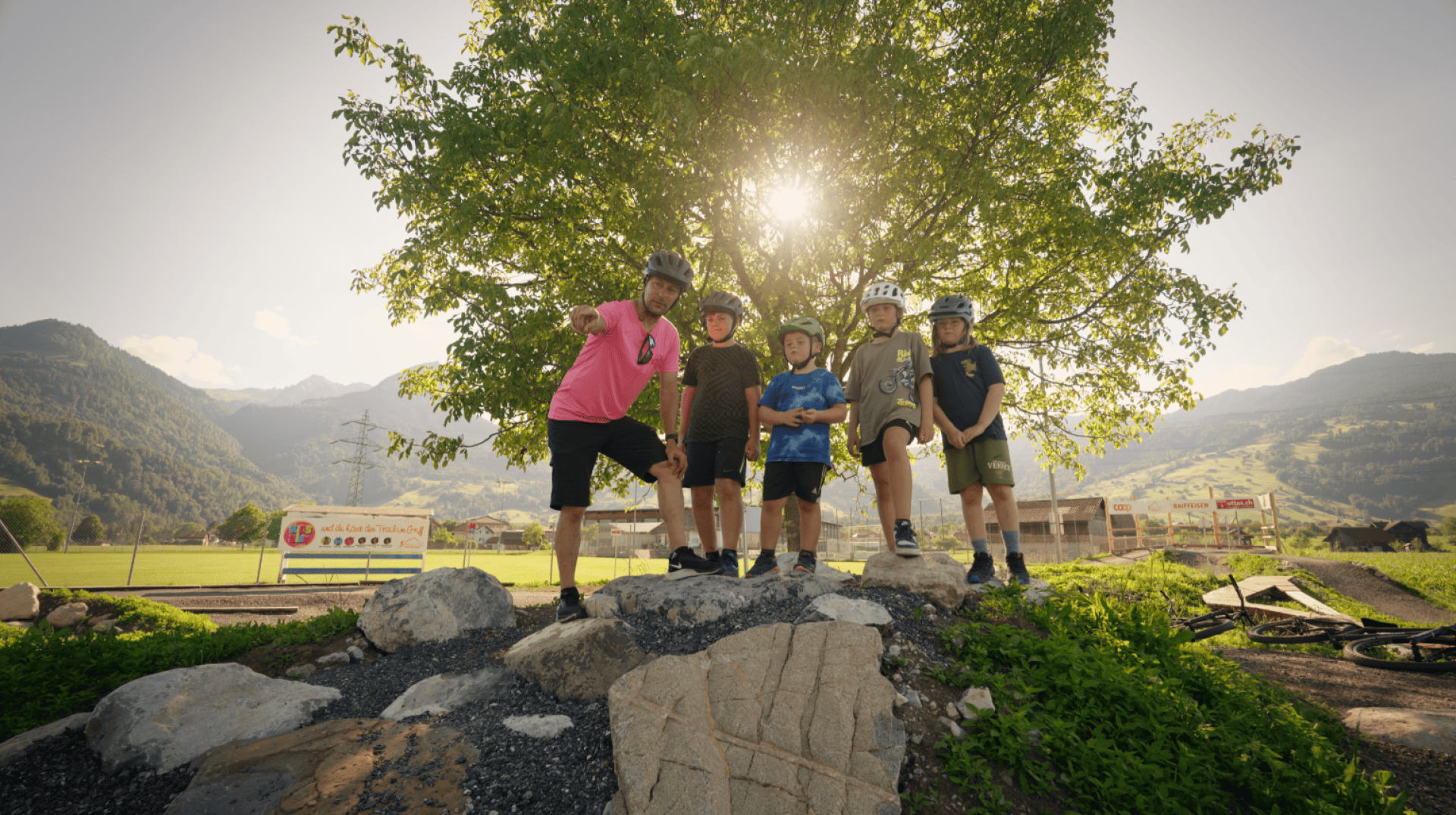 Vier Kinder mit Helmen und ein Mann in einem rosa Hemd posieren auf Felsen vor einem Baum, durch dessen Blätter die Sonne scheint. Im Hintergrund sind Berge und Felder zu sehen, die eine malerische Kulisse bilden.