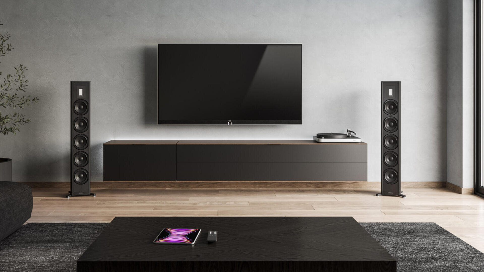 Ein modernes Wohnzimmer mit einem an der Wand montierten Flachbildfernseher, zwei hohen schwarzen Lautsprechern, einem Plattenspieler auf einem niedrigen Schrank, einem Couchtisch mit einem Tablet und einer Fernbedienung sowie einem hellen Holzboden.