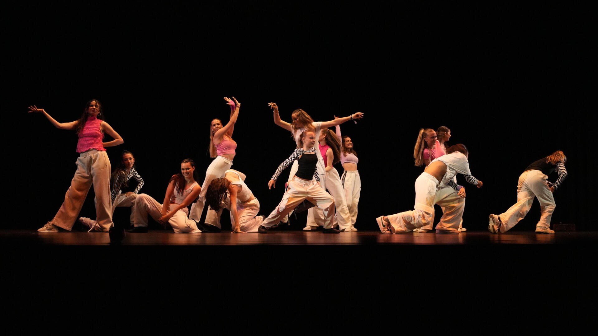 Eine Gruppe von Tänzern auf der Bühne in dramatischen Posen, in weiten beigen Hosen und bunten Oberteilen, die im Scheinwerferlicht vor einem dunklen Hintergrund auftreten.