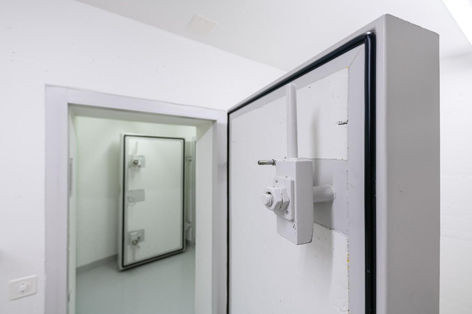 Eine weiße, verstärkte Metalltür mit einem Schwerlastschloss ist leicht geöffnet und führt in einen Raum mit einer weiteren, ähnlich gesicherten Tür im Hintergrund. Der Raum wirkt sauber, hell und industriell.