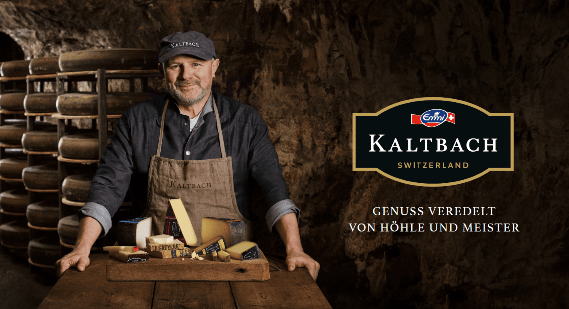 Ein lächelnder Mann mit Mütze und Schürze steht in einer Käsehöhle und präsentiert ein Holztablett mit verschiedenen Käsesorten. Hinter ihm stehen Regale mit reifenden Käselaiben. Der Text lautet: "Kaltbach Schweiz. Genuss veredelt von Höhle und Meister.".
