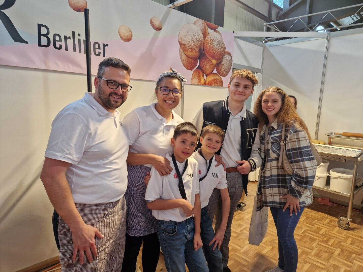 Sechs Personen, darunter zwei Erwachsene und vier Kinder, stehen lächelnd bei einer Indoor-Veranstaltung zusammen. Einige tragen weiße Hemden und Schürzen. Hinter ihnen ist ein Banner mit Bildern von Gebäck und dem Wort Berliner zu sehen.