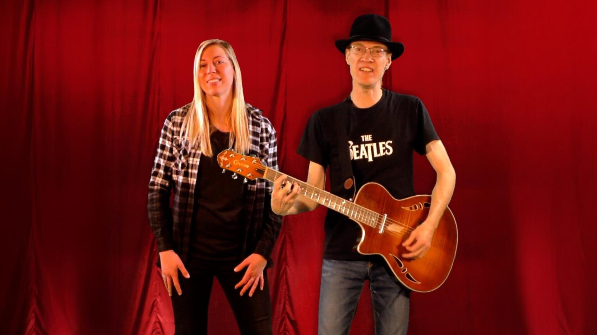 Zwei Personen stehen vor einem roten Vorhang; die eine lächelt, die andere trägt einen Hut und eine Brille, spielt eine akustische Gitarre und trägt ein T-Shirt der Beatles.