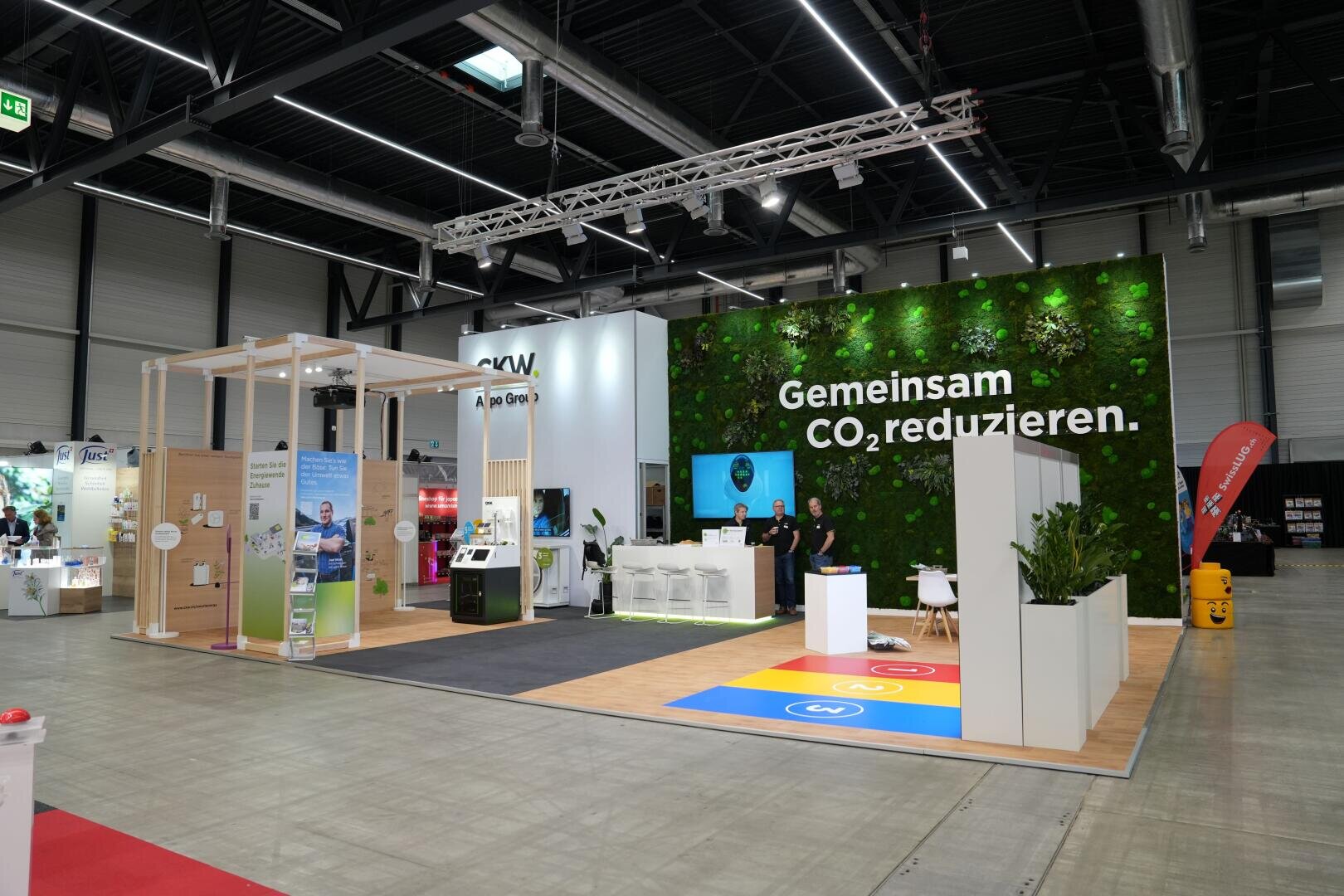 Ein moderner Messestand mit einer grünen Pflanzenwand, die den deutschen Text Gemeinsam CO₂ reduzieren zeigt. Zwei Personen stehen an einem Empfangstresen; der Stand zeichnet sich durch umweltfreundliche Displays und einen offenen, gut beleuchteten Raum aus.