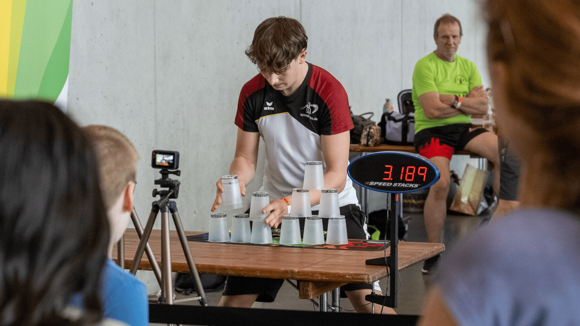 Ein Jugendlicher stapelt schnell Becher an einem Wettbewerbstisch und wird dabei von einer kleinen Kamera gefilmt. Ein digitaler Timer zeigt 3,189 Sekunden an. Im Hintergrund schauen Zuschauer zu.