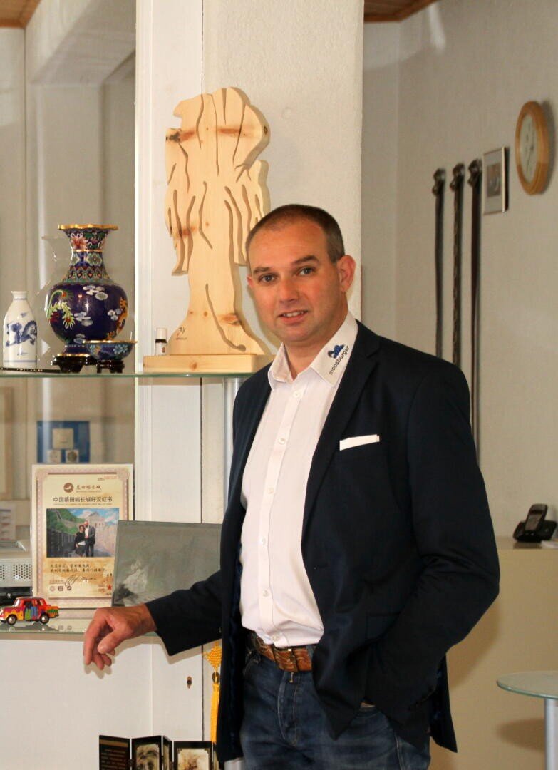 Ein Mann in einem marineblauen Blazer und weißem Hemd steht in einem Haus neben einem Regal mit Dekorationsartikeln, einer Holzskulptur, einer bunten Vase und gerahmten Urkunden und lächelt leicht in die Kamera.