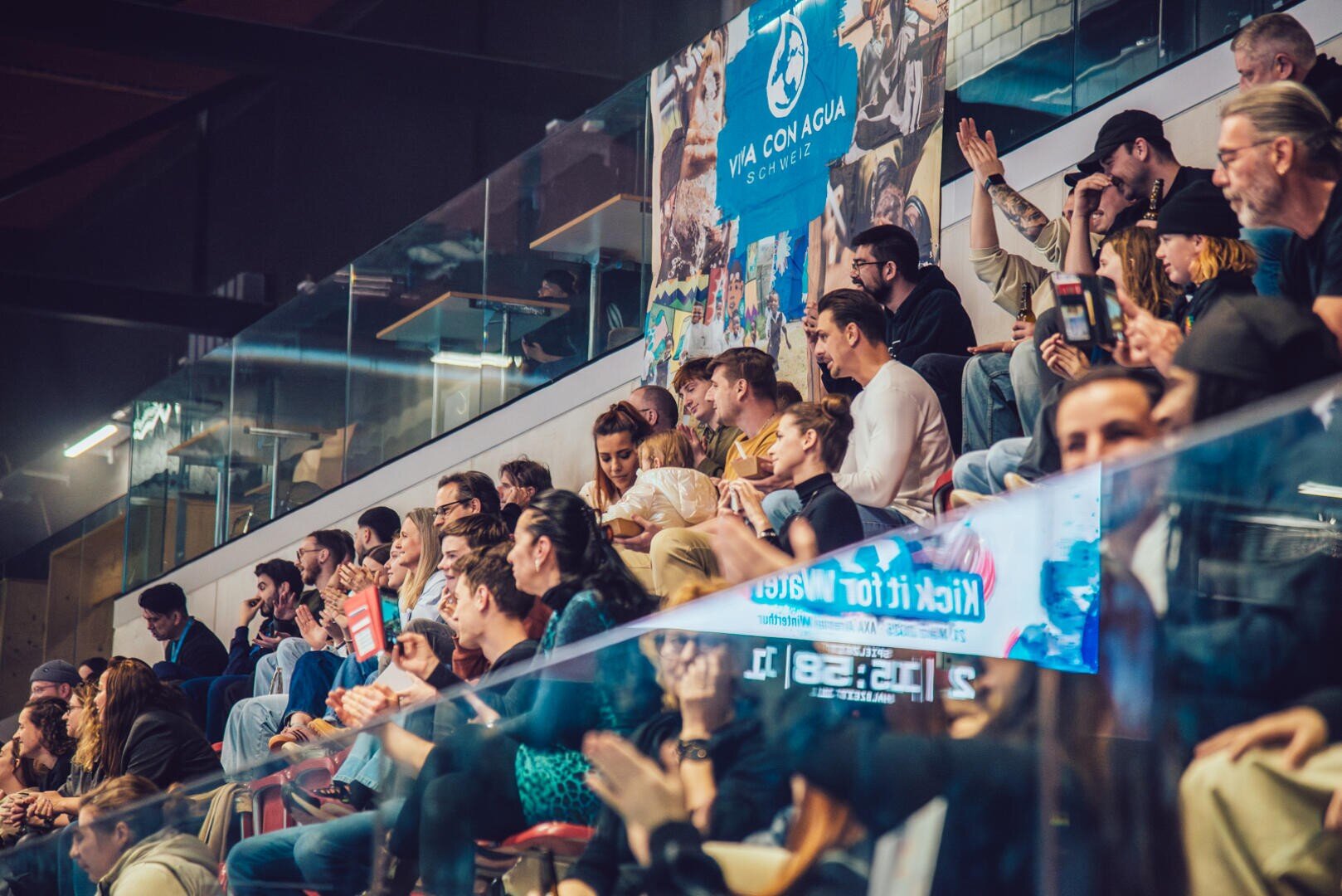 Eine bunt gemischte Gruppe von Menschen sitzt auf abgestuften Sitzplätzen bei einer Indoor-Veranstaltung und schaut aufmerksam zu. Einige klatschen, andere unterhalten sich oder benutzen ihre Telefone. Im Hintergrund ist ein großes Transparent für "Viva con Agua" zu sehen.