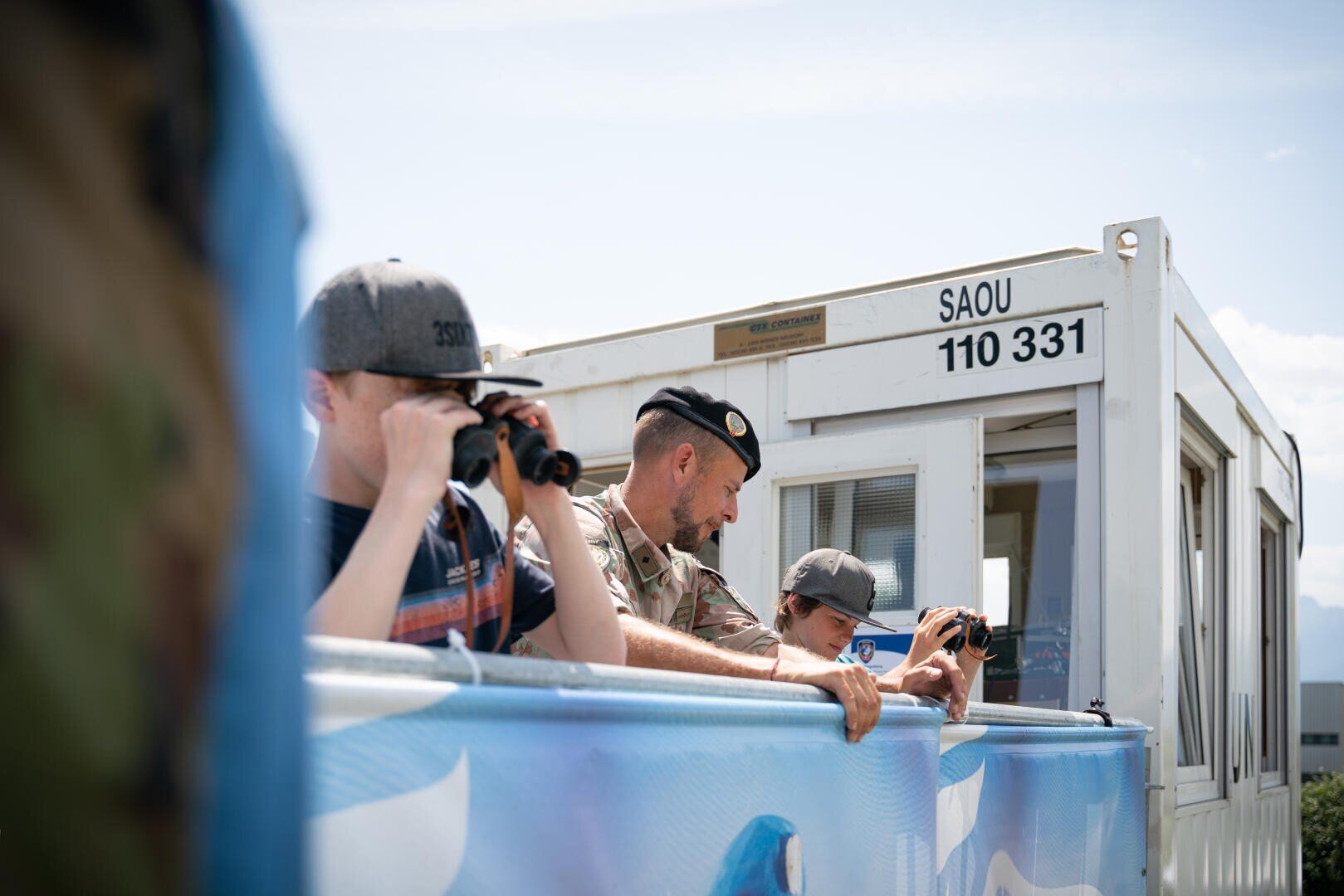 Drei Personen, zwei in Freizeitkleidung mit Ferngläsern und eine in Militäruniform, stehen an einer blauen Absperrung in der Nähe eines weißen Gebäudes mit der Aufschrift SAOU 110 331 unter strahlendem Himmel.