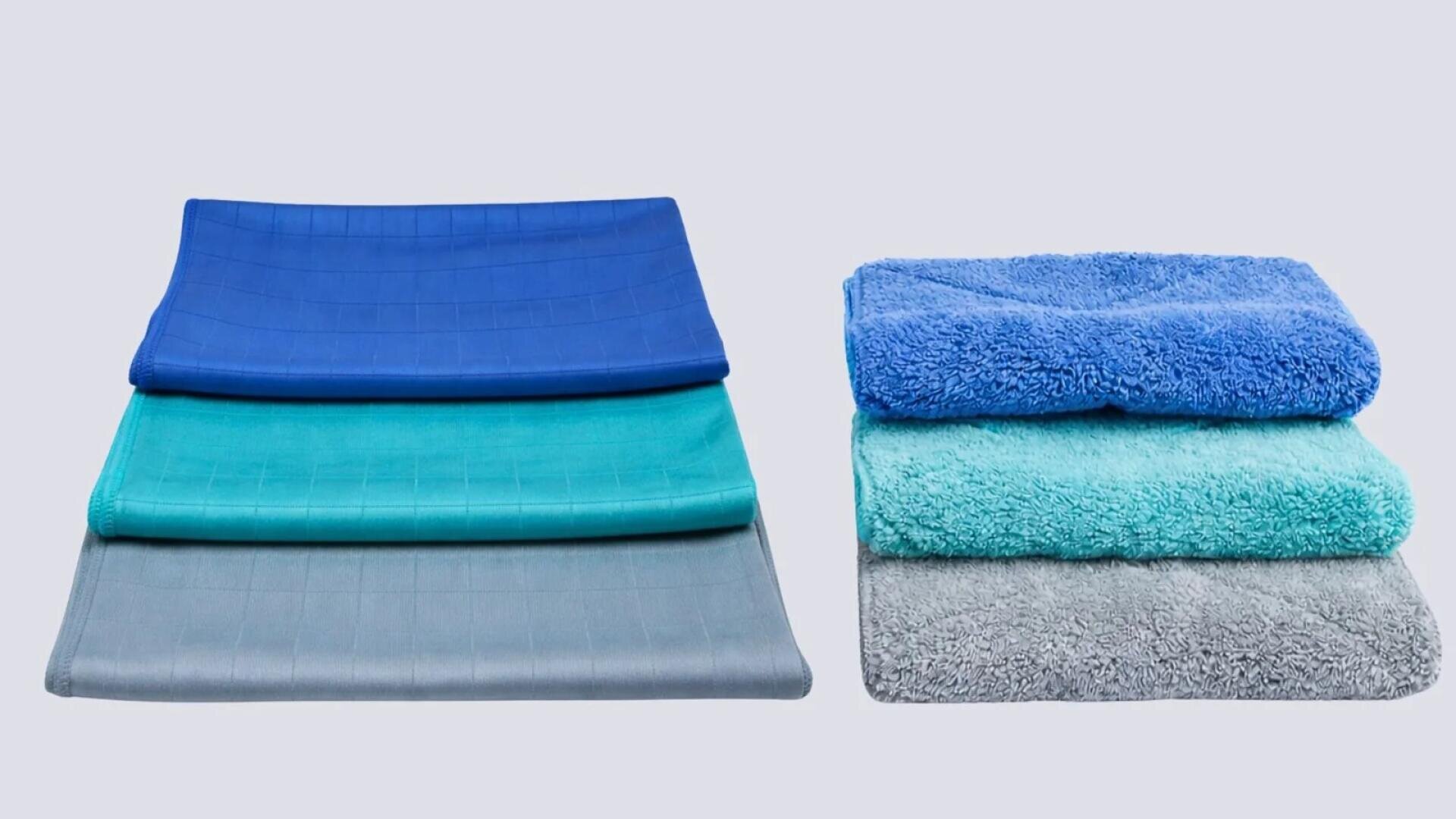 Drei dünne, glatte Handtücher in Blau, Türkis und Grau sind auf der linken Seite gestapelt. Drei flauschige, strukturierte Handtücher in ähnlichen Farben sind auf der rechten Seite gestapelt, alle vor einem hellgrauen Hintergrund.