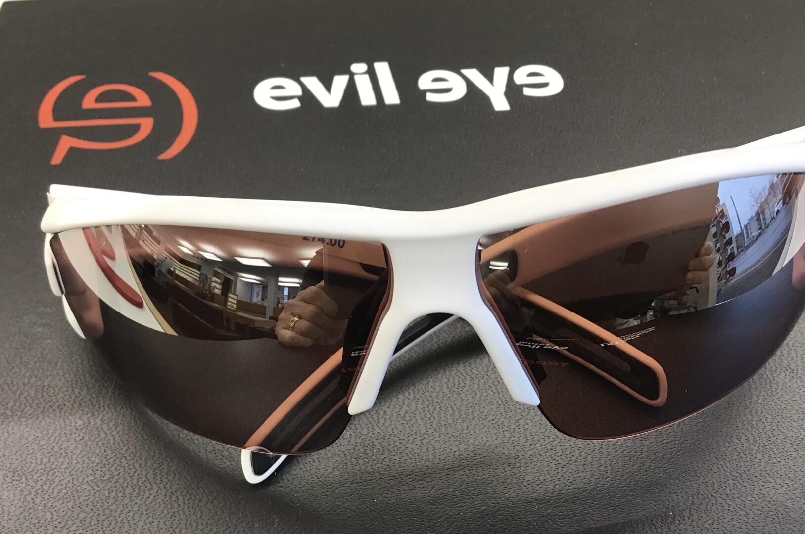 Eine weiße Sport-Sonnenbrille mit verspiegelten Gläsern sitzt auf einer schwarzen Fläche mit dem Logo und Text des Bösen Auges. In den Gläsern spiegeln sich eine Hand, die ein Telefon hält, und einige Innenräume.