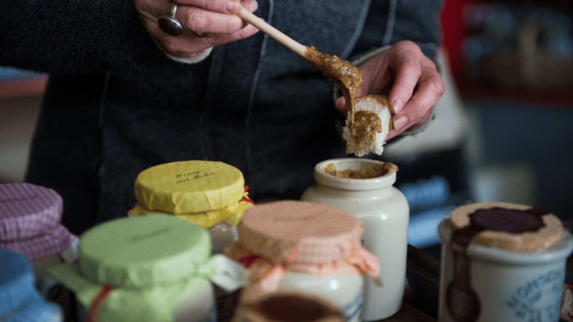 Eine Person streicht mit einem Holzstäbchen eine braune Paste auf ein Stück Brot, während vor ihr auf dem Tisch mehrere Gläser mit bunten Stoffdeckeln stehen.