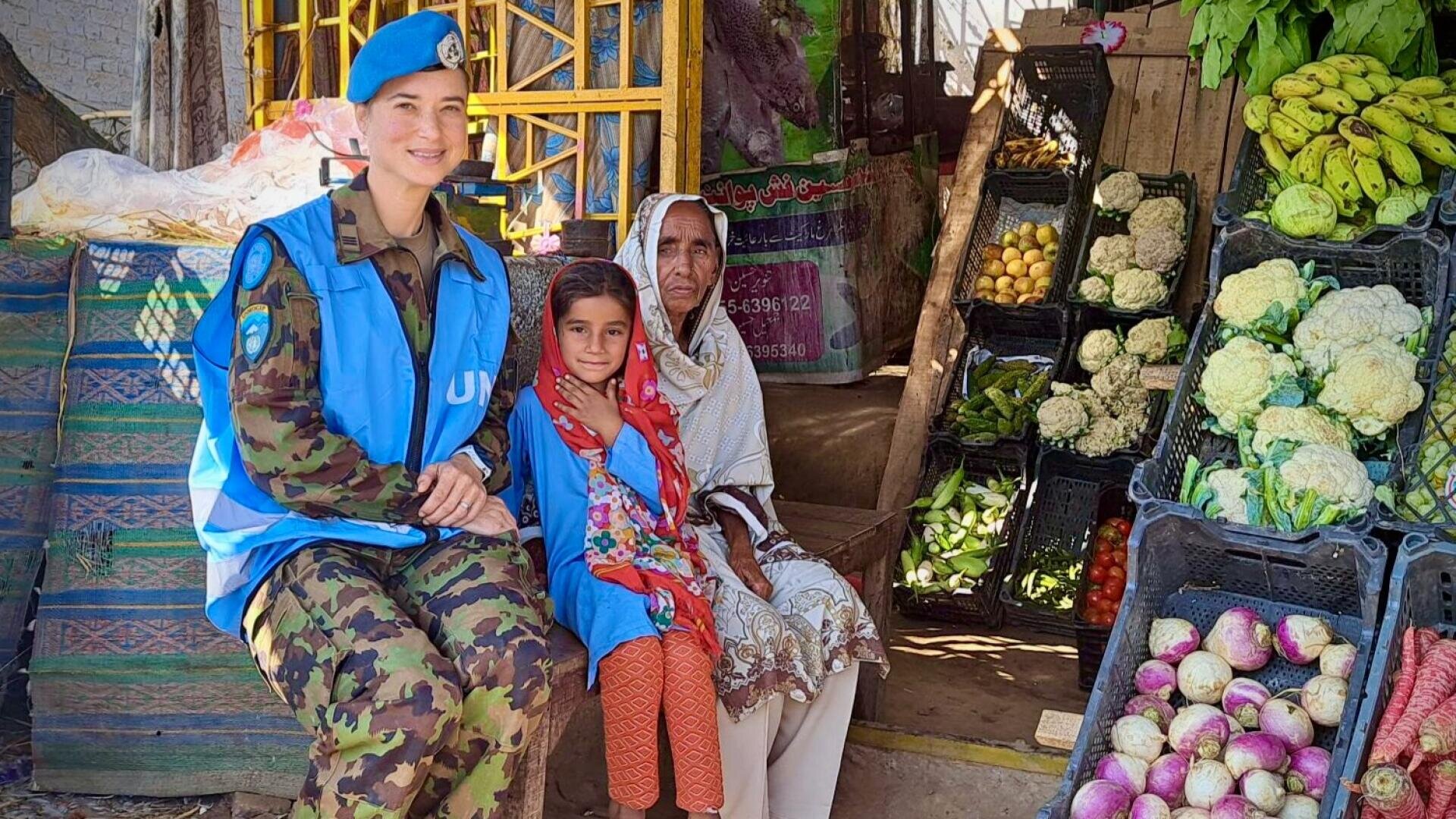 Eine UN-Friedenswächterin in Uniform sitzt lächelnd neben einer älteren Frau und einem jungen Mädchen an einem Gemüsestand, an dem verschiedene Gemüsesorten wie Blumenkohl, Rüben und Bananen in Kisten angeboten werden.