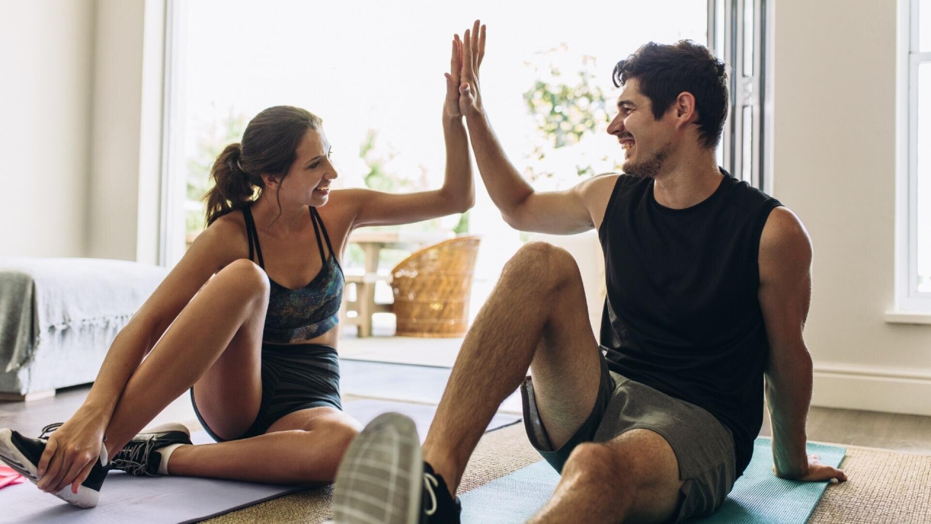 Eine Frau und ein Mann in Trainingskleidung sitzen drinnen auf Yogamatten, lächeln sich an und geben sich nach dem gemeinsamen Training ein High Five. Das Sonnenlicht fällt durch eine offene Tür im Hintergrund.