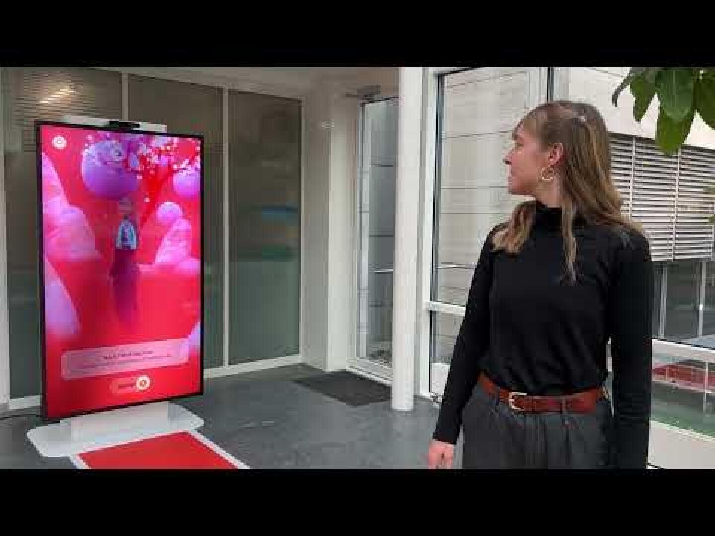 Eine Frau steht in einem Innenraum und blickt auf einen großen vertikalen Digitalbildschirm, auf dem ein lebhaftes, farbenfrohes Bild und Text zu sehen ist. Der Bildschirm befindet sich in der Nähe von Glastüren und Fenstern, und auf dem Boden davor liegt ein roter Teppich.