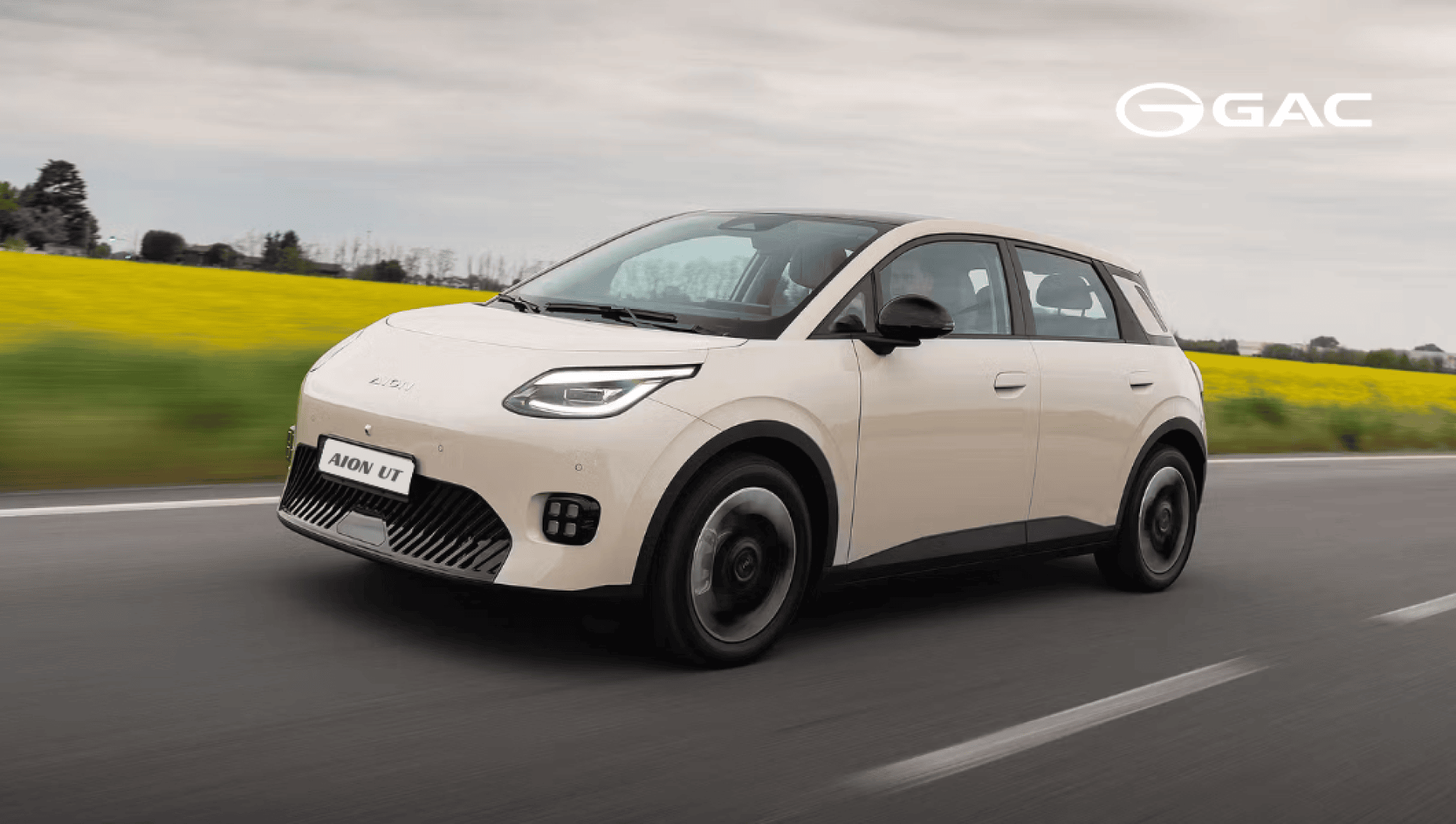 Ein cremefarbenes GAC AION-Elektroauto fährt auf einer Straße vorbei an einem gelben Feld und einem bewölkten Himmel, mit dem GAC-Logo in der oberen rechten Ecke.