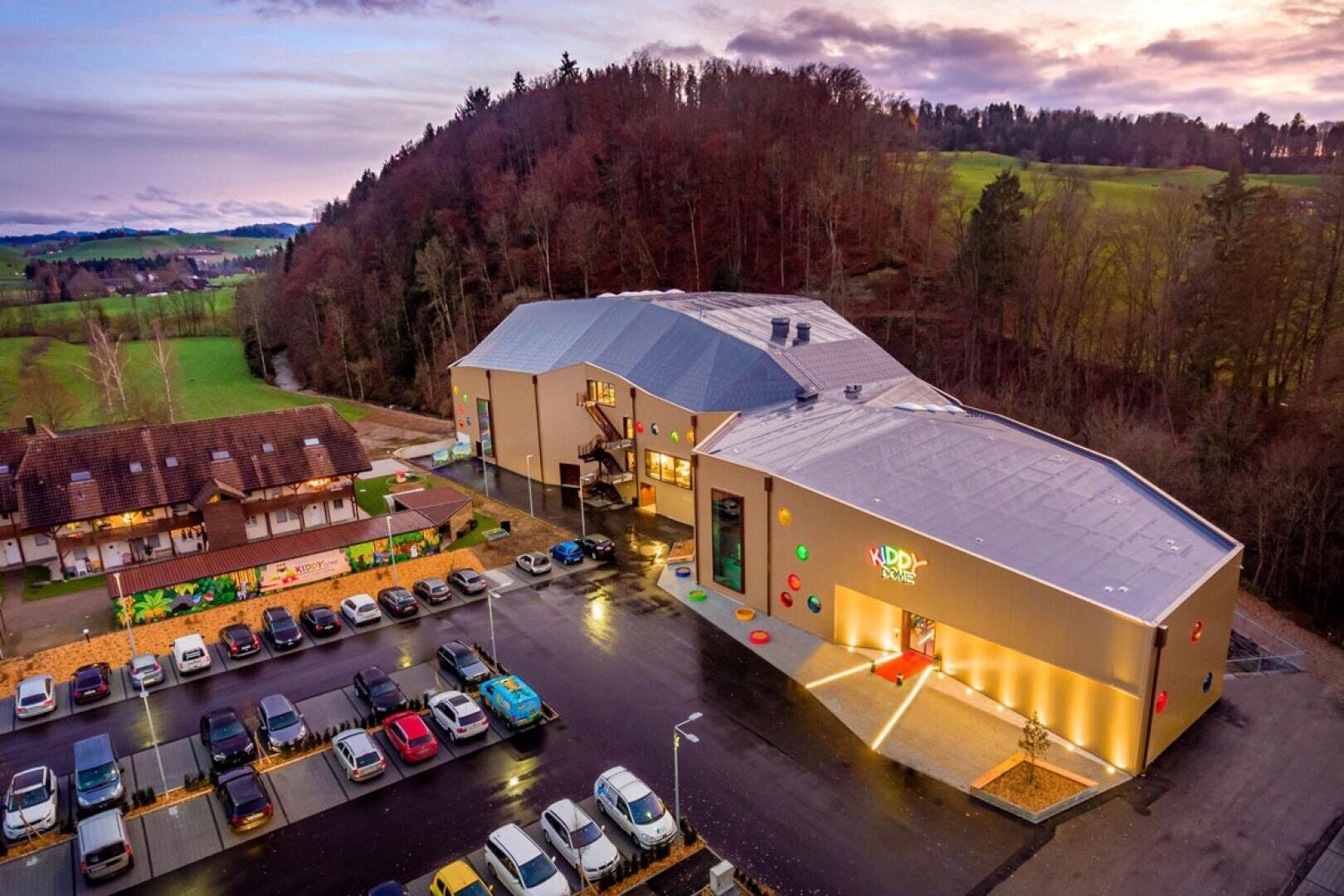 Luftaufnahme eines modernen Freizeitzentrums mit Kiddy-Dome-Beschilderung, bunten Akzenten und einem großen Parkplatz, umgeben von Bäumen, Wiesen und Hügeln bei Sonnenuntergang.