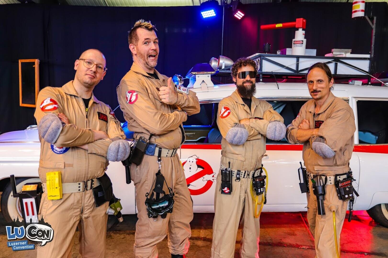 Vier als Ghostbusters verkleidete Personen posieren selbstbewusst vor einem weißen Ghostbusters-Auto auf einem Kongress. Sie tragen braune Overalls mit Ghostbusters-Aufnähern und Gürteln. In der Ecke ist das Lucon-Logo zu sehen.