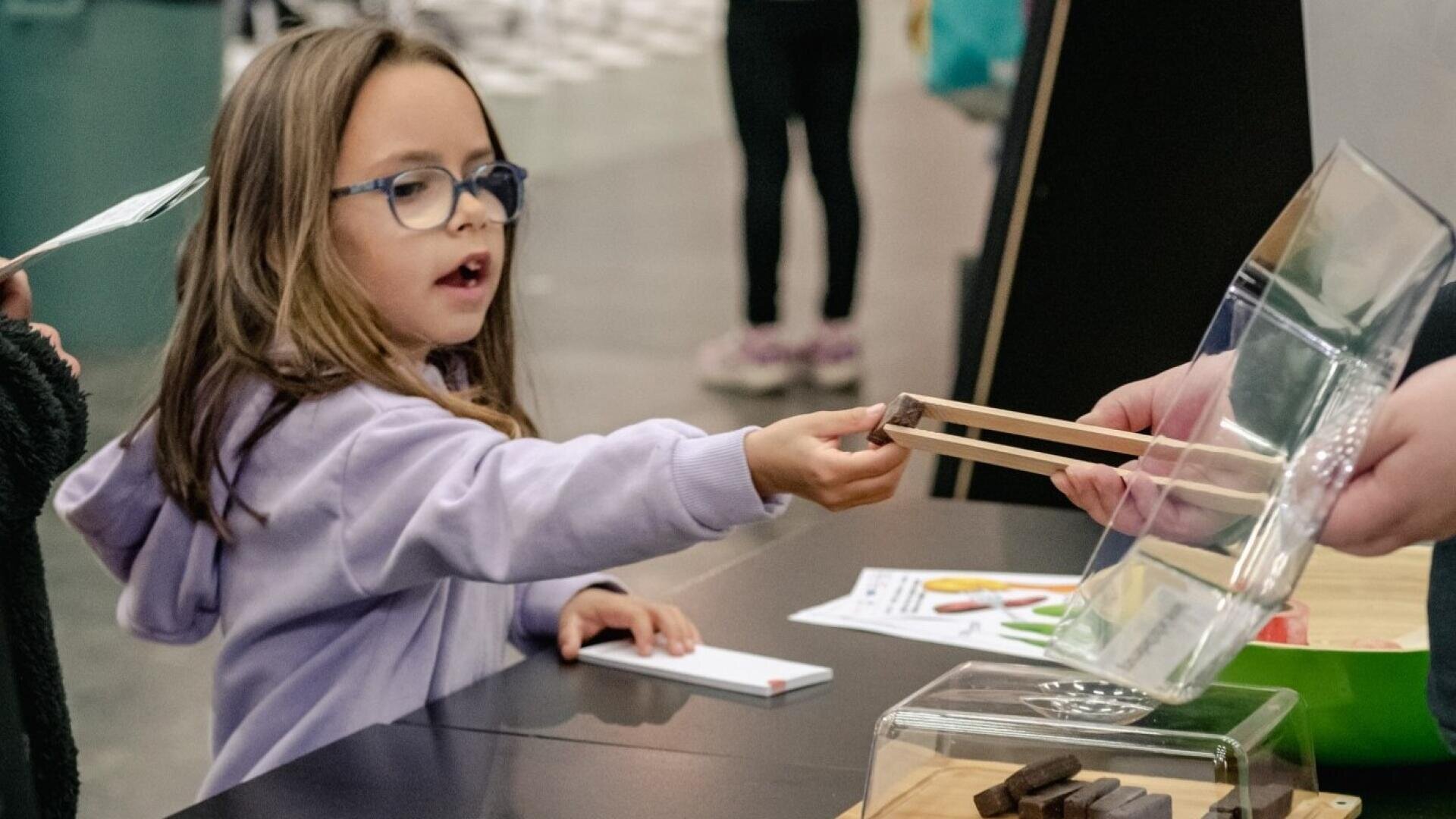 Ein junges Mädchen mit Brille und lilafarbenem Kapuzenpulli streckt die Hand aus, als ihr jemand an einem Ausstellungstisch ein Stück Essen mit einer Zange reicht. Sie sieht neugierig und aufmerksam aus. Im Hintergrund sind weitere Personen zu sehen.