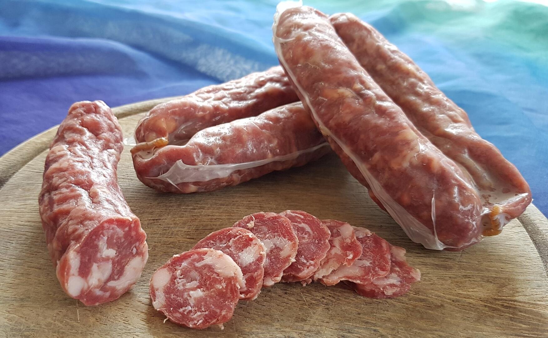 Mehrere ganze und in Scheiben geschnittene gepökelte Würste sind auf einem Holzbrett angeordnet, mit einem bunten Tuch im Hintergrund. Die Würste erscheinen mit Fett marmoriert und haben eine rötliche Farbe.