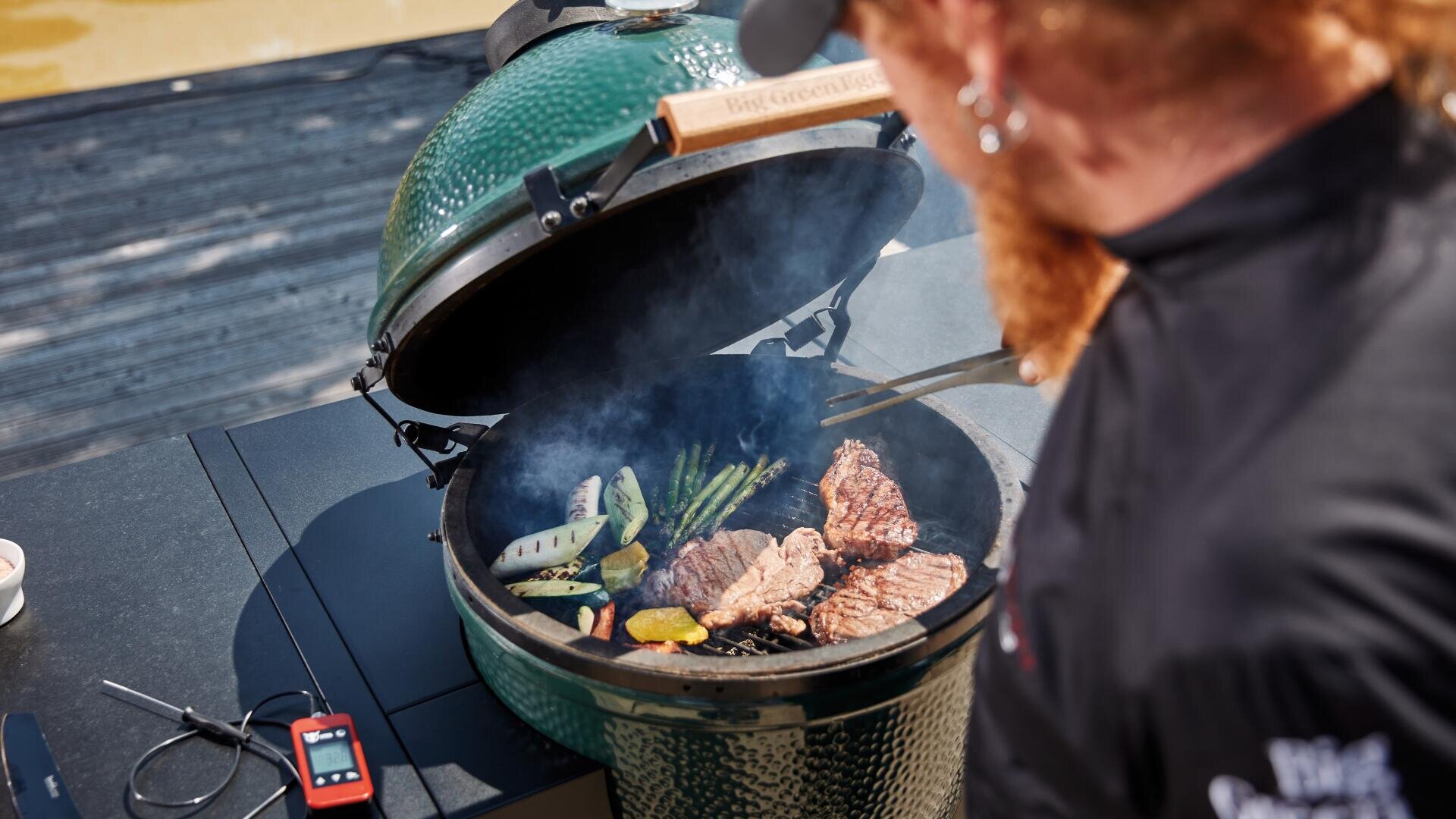 Eine Person grillt Steaks, Spargel und anderes Gemüse auf einem grünen Keramikgrill im Freien. Rauch steigt vom Grill auf, und ein Fleischthermometer liegt auf dem Tresen daneben.