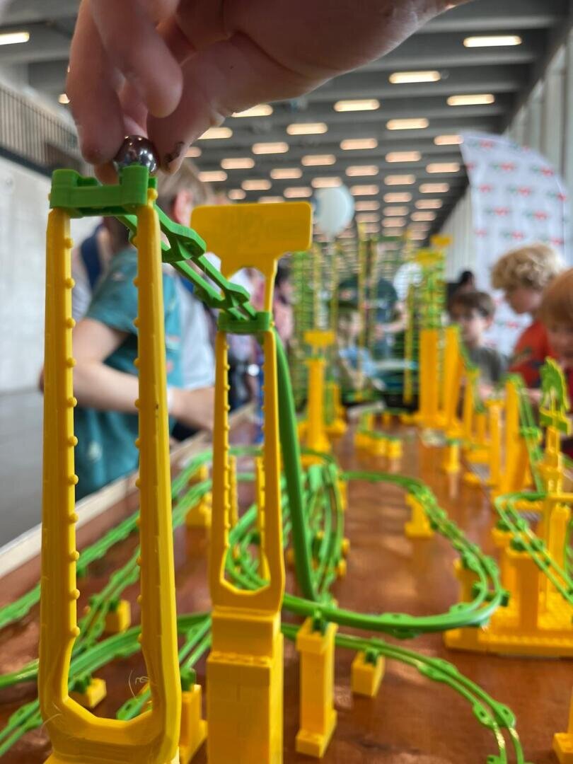 Nahaufnahme einer Hand, die eine Murmel auf eine leuchtend gelbe und grüne Kunststoffmurmelbahn legt, wobei Kinder und bunte Strukturen im unscharfen Hintergrund zu sehen sind.