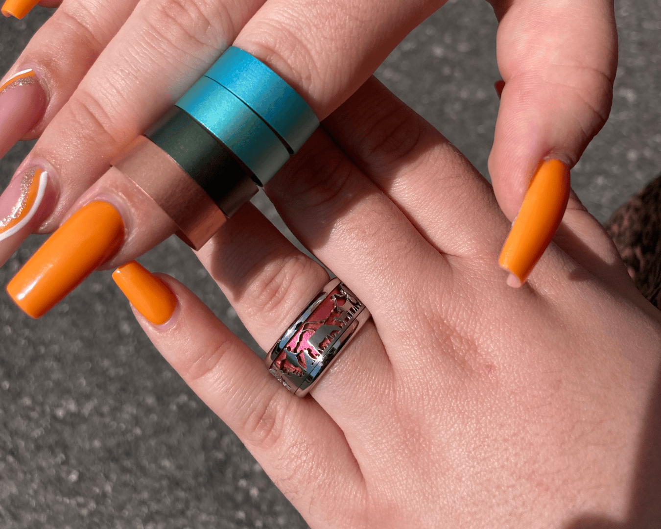 Zwei Hände mit langen, leuchtend orangefarbenen Nägeln; eine Hand trägt einen silbernen Ring mit einem rosafarbenen Muster, während die andere vier metallische Ringe in Blau, Petrol, Grün und Kupfer an den Fingern trägt. Der Hintergrund besteht aus grauem Pflaster.