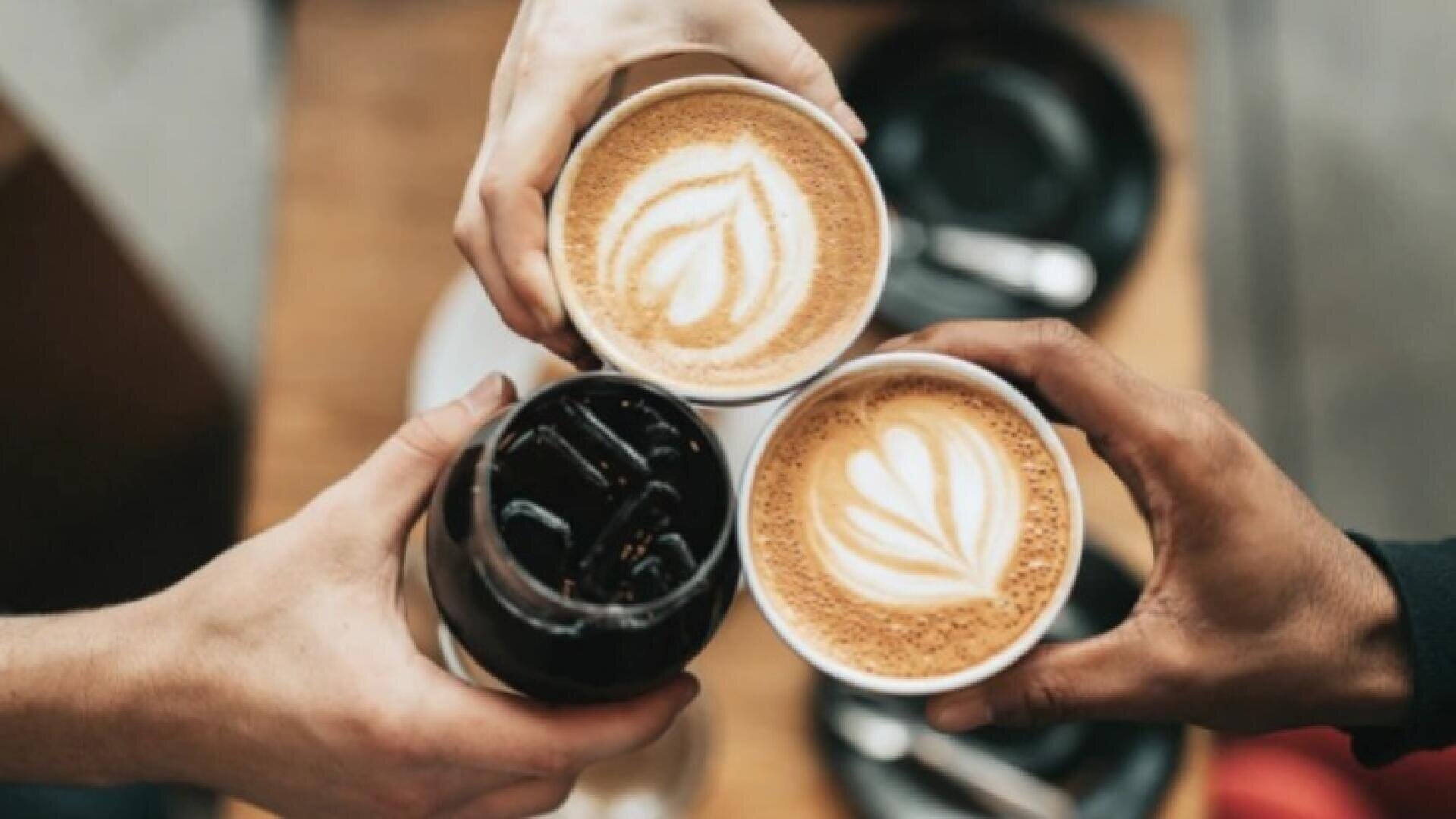 Drei Personen stoßen über einem Holztisch mit Getränken an, zwei mit Latte Art und eine mit einem Glas Eiskaffee.