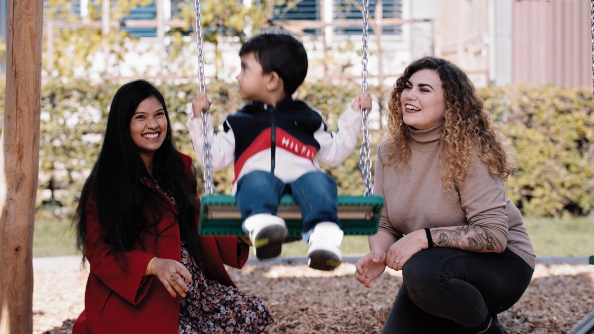 Zwei Frauen lächeln und knien neben einem kleinen Jungen, der auf einer Schaukel auf einem Spielplatz sitzt. Der Junge ist in Bewegung und wendet sich ab, während die Frauen glücklich und mit ihm beschäftigt aussehen. Im Hintergrund sind Bäume und ein Gebäude zu sehen.