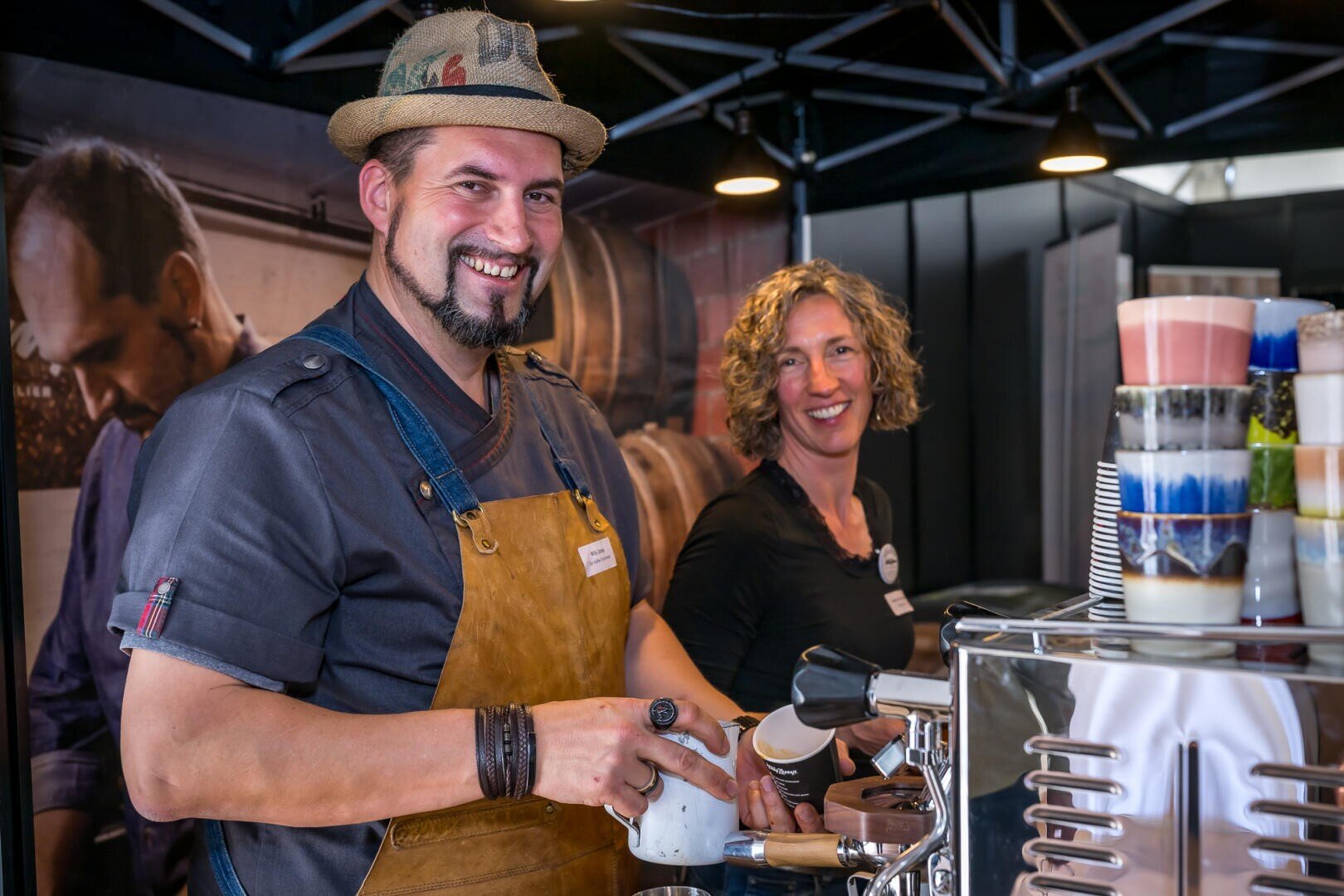Ein lächelnder Mann mit brauner Schürze und Hut steht an einer Kaffeemaschine, die Milch aufschäumt, während eine Frau mit lockigem Haar neben ihm lächelt. Auf dem Tresen sind Stapel von bunten Tassen zu sehen. Beide scheinen an einem Kaffeestand zu arbeiten.