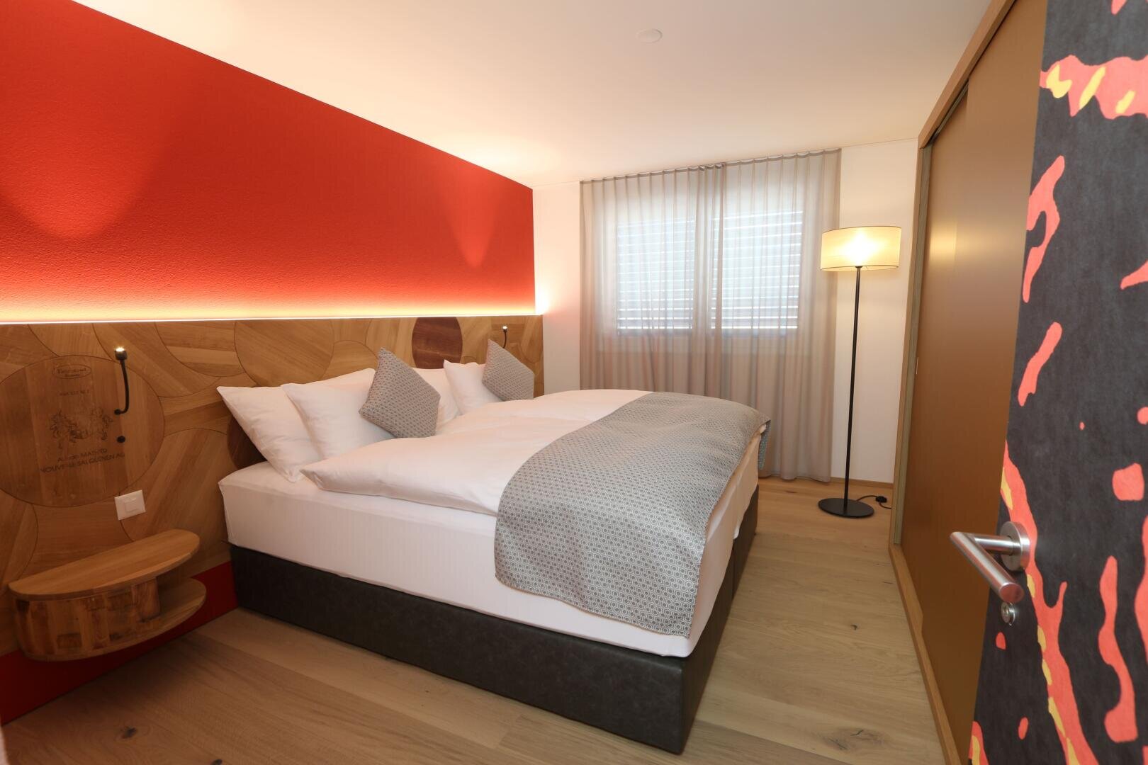 Ein modernes Hotelzimmer mit einem Doppelbett, grauer Bettwäsche, roter Akzentwand, hölzernem Kopfteil, Nachttischen, Stehlampe, durchsichtigen Vorhängen vor den Fenstern und hellem Holzfußboden.
