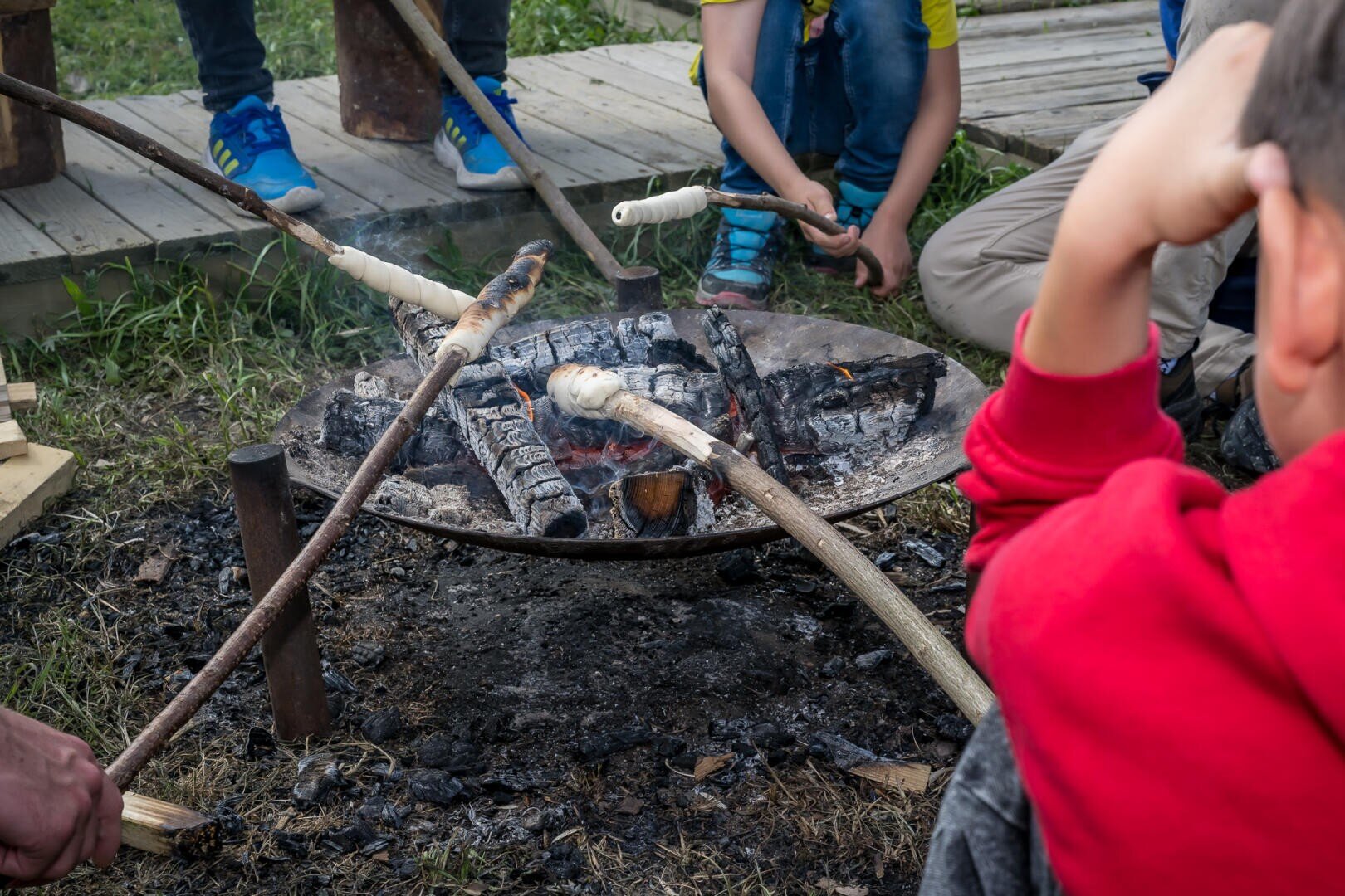 Kinder sitzen um eine Feuerstelle herum und rösten Brotteig auf Stöcken über heißen Kohlen im Freien. Der Boden ist verkohlt, und die Szene scheint Teil eines Lagers oder einer Aktivität im Freien zu sein.