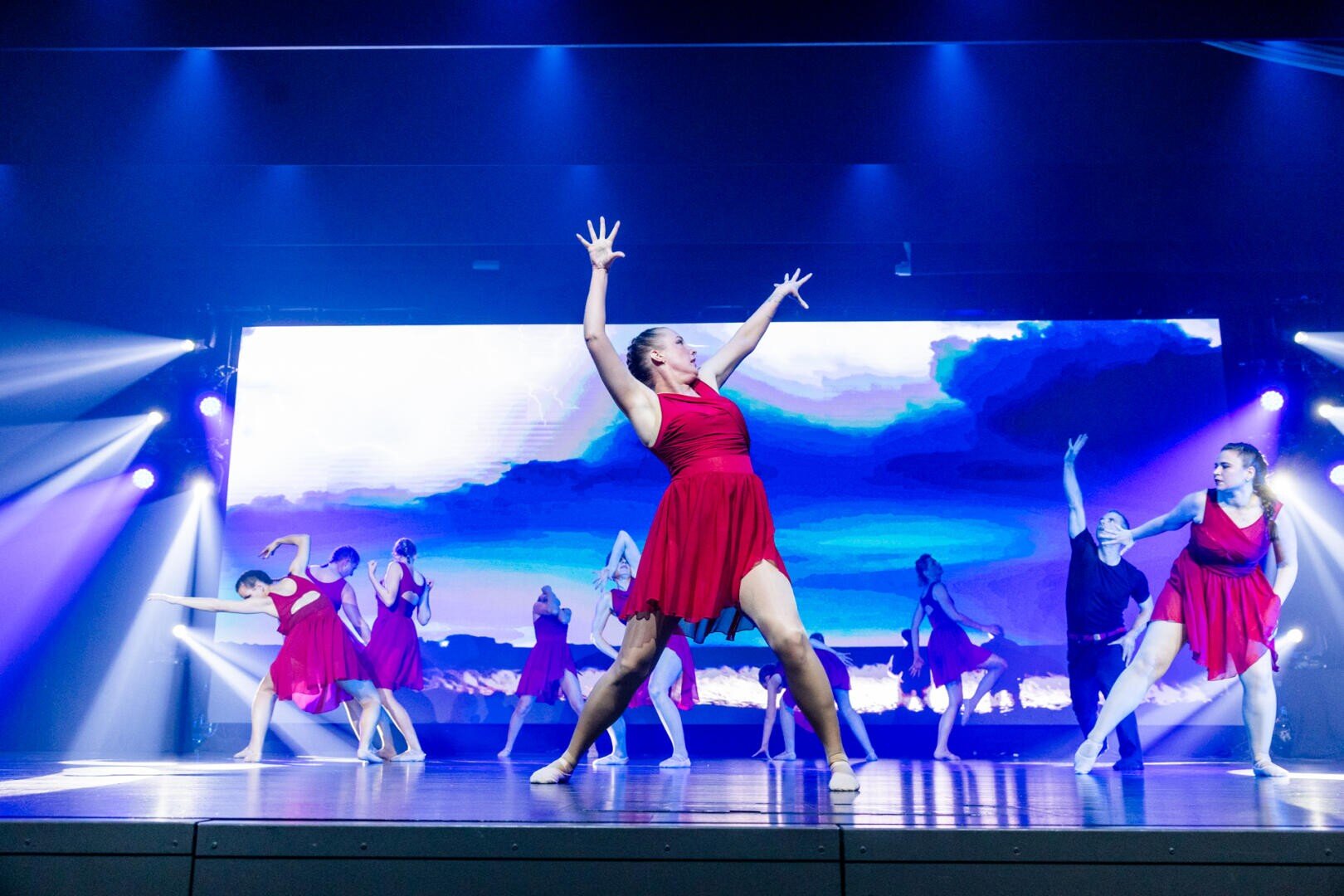 Eine Gruppe von Tänzerinnen und Tänzern in roten Kleidern führt auf der Bühne vor dramatischer Beleuchtung und einem blauen, wolkenähnlichen digitalen Hintergrund ein ausdrucksstarkes Programm auf.