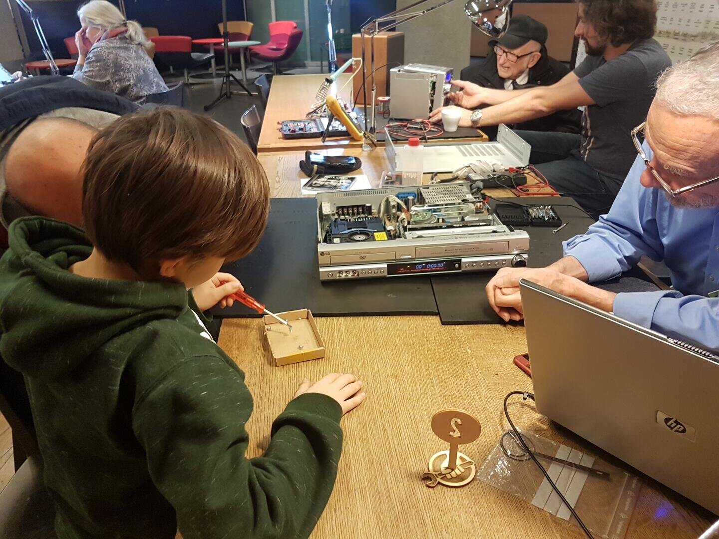Ein Kind arbeitet an einem Tisch mit einem Schraubenzieher an einem kleinen Elektronikprojekt, während mehrere Erwachsene in der Nähe in einer Werkstatt an anderen elektronischen Geräten arbeiten und dabei helfen.