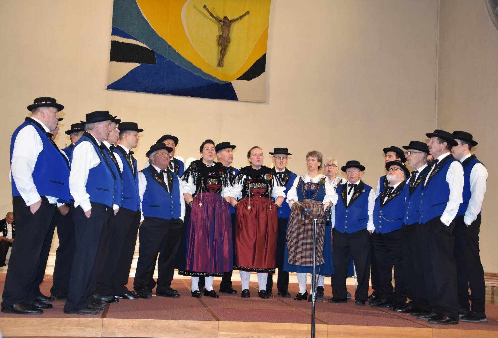 Ein Chor von Männern und Frauen in traditioneller bayerischer Kleidung singt gemeinsam auf einer hölzernen Bühne in einer Kirche, hinter der ein bunter religiöser Wandteppich hängt.