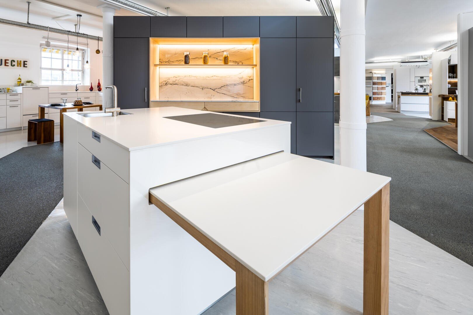 Moderne Küche mit weißer Insel, eingebautem Kochfeld, ausziehbarer Arbeitsplattenerweiterung mit Holzbeinen, dunkelgrauen Schränken und beleuchteten Regalen mit Gläsern vor einer Marmorabdeckung. Geräumig, offenes Layout mit reichlich natürlichem Licht.