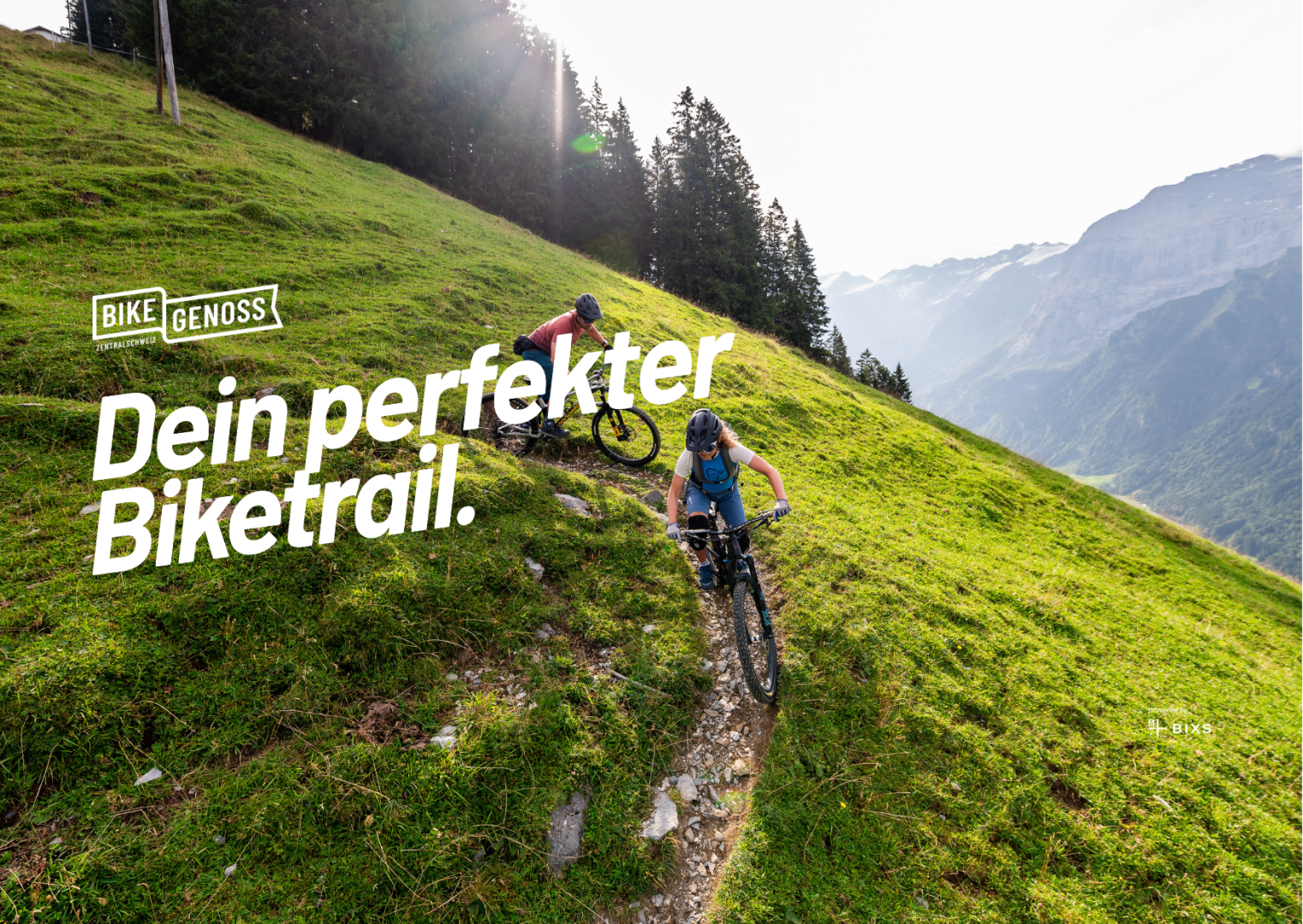 Zwei Personen fahren mit dem Mountainbike auf einem schmalen Pfad entlang eines grasbewachsenen Hangs mit Bäumen, Bergen und einem hellen Himmel im Hintergrund. Der deutsche Text lautet: "Dein perfekter Biketrail.".