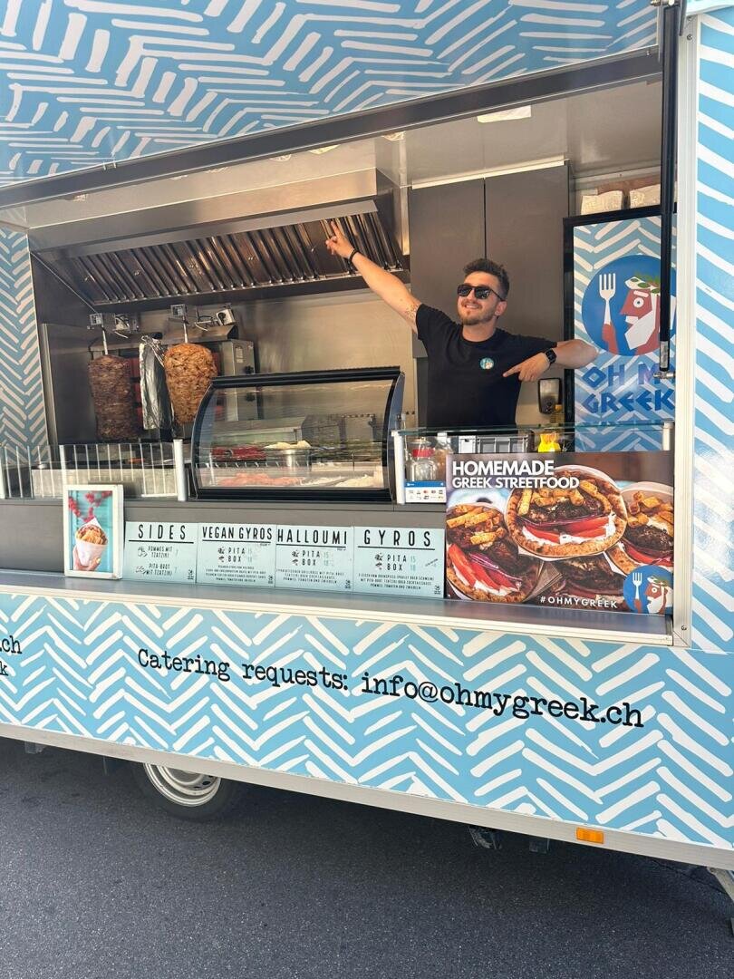 Ein Mann mit Sonnenbrille steht lächelnd und winkend am Schaufenster eines blauen Imbisswagens mit griechischem Motiv. Im Inneren des Wagens sind Schawarma-Spieße, eine Speisekarte und Lebensmittelauslagen zu sehen.
