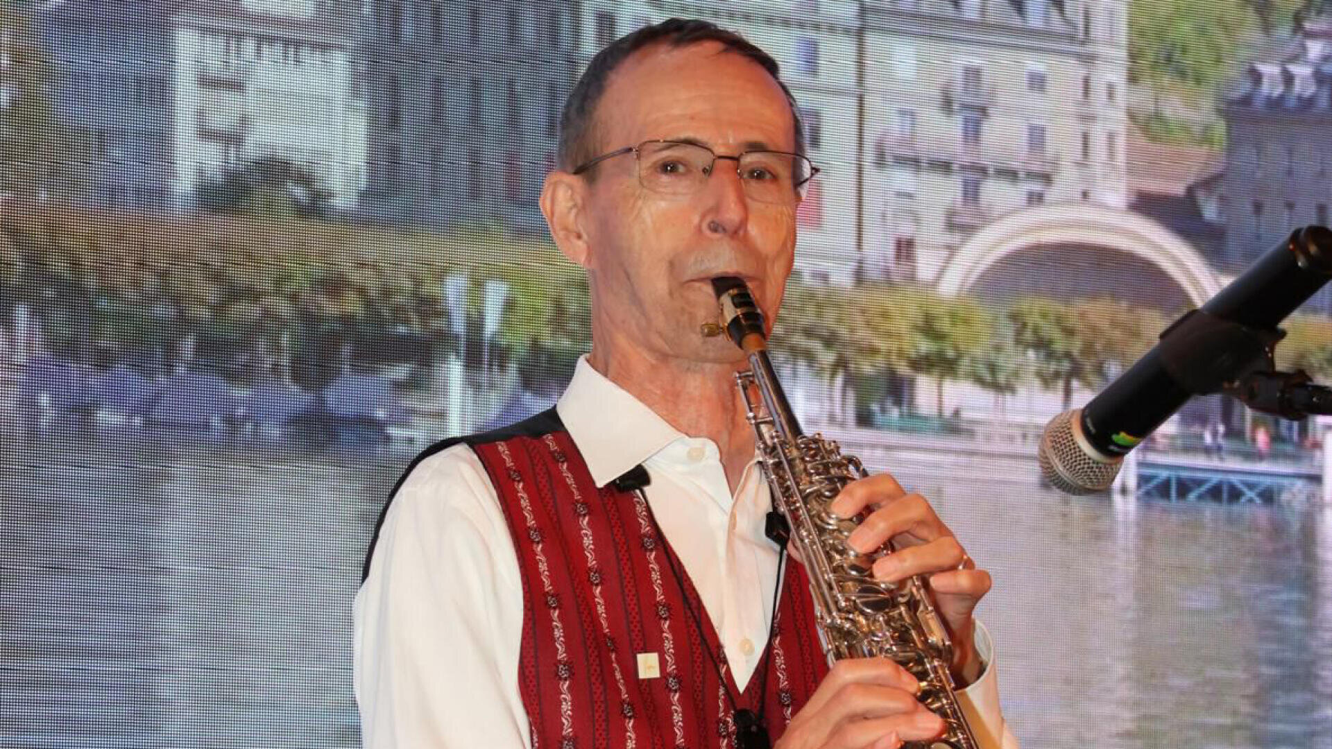 Ein älterer Mann mit Brille und rot gemusterter Weste spielt auf der Bühne Saxophon, in der Nähe befindet sich ein Mikrofon, im Hintergrund eine malerische Stadtkulisse.
