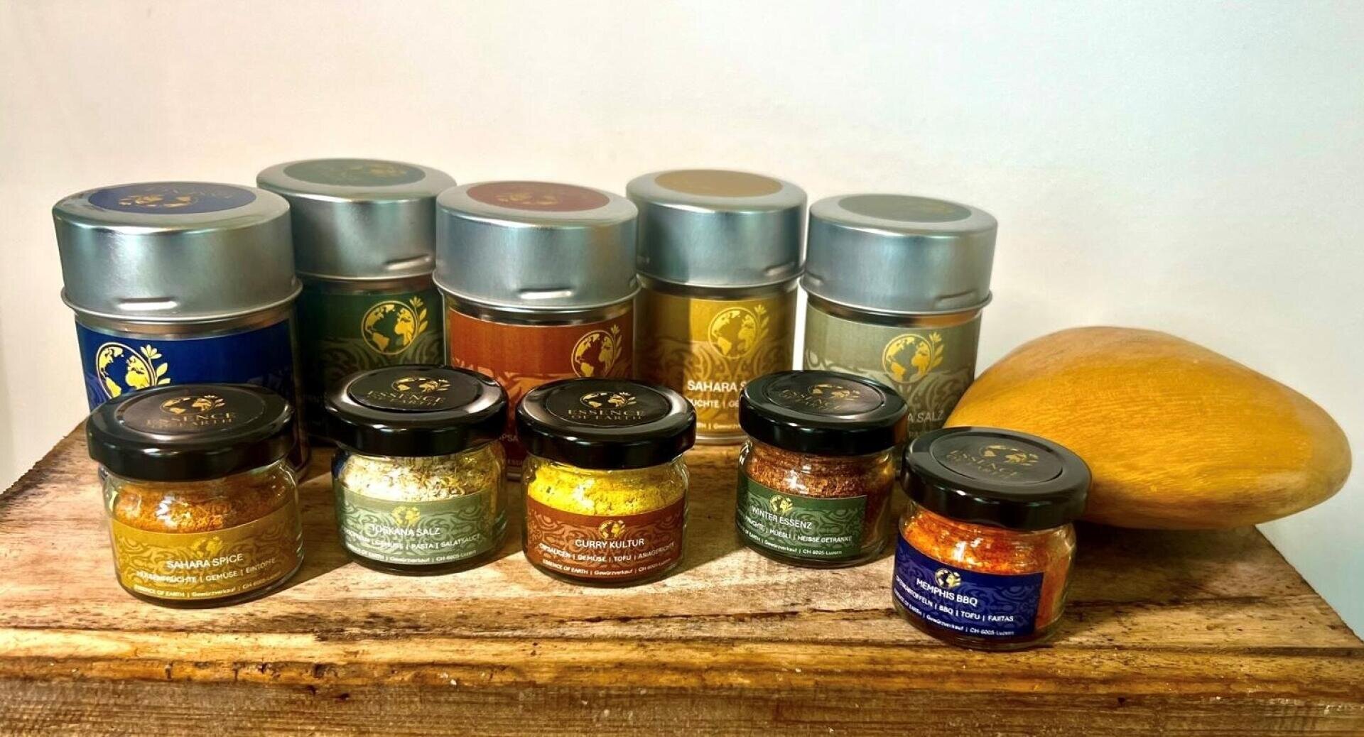 Eine Reihe von Gewürzgläsern und Dosen mit bunten Etiketten sind auf einer Holzfläche neben einer reifen gelben Mango vor einem schlichten Hintergrund angeordnet.