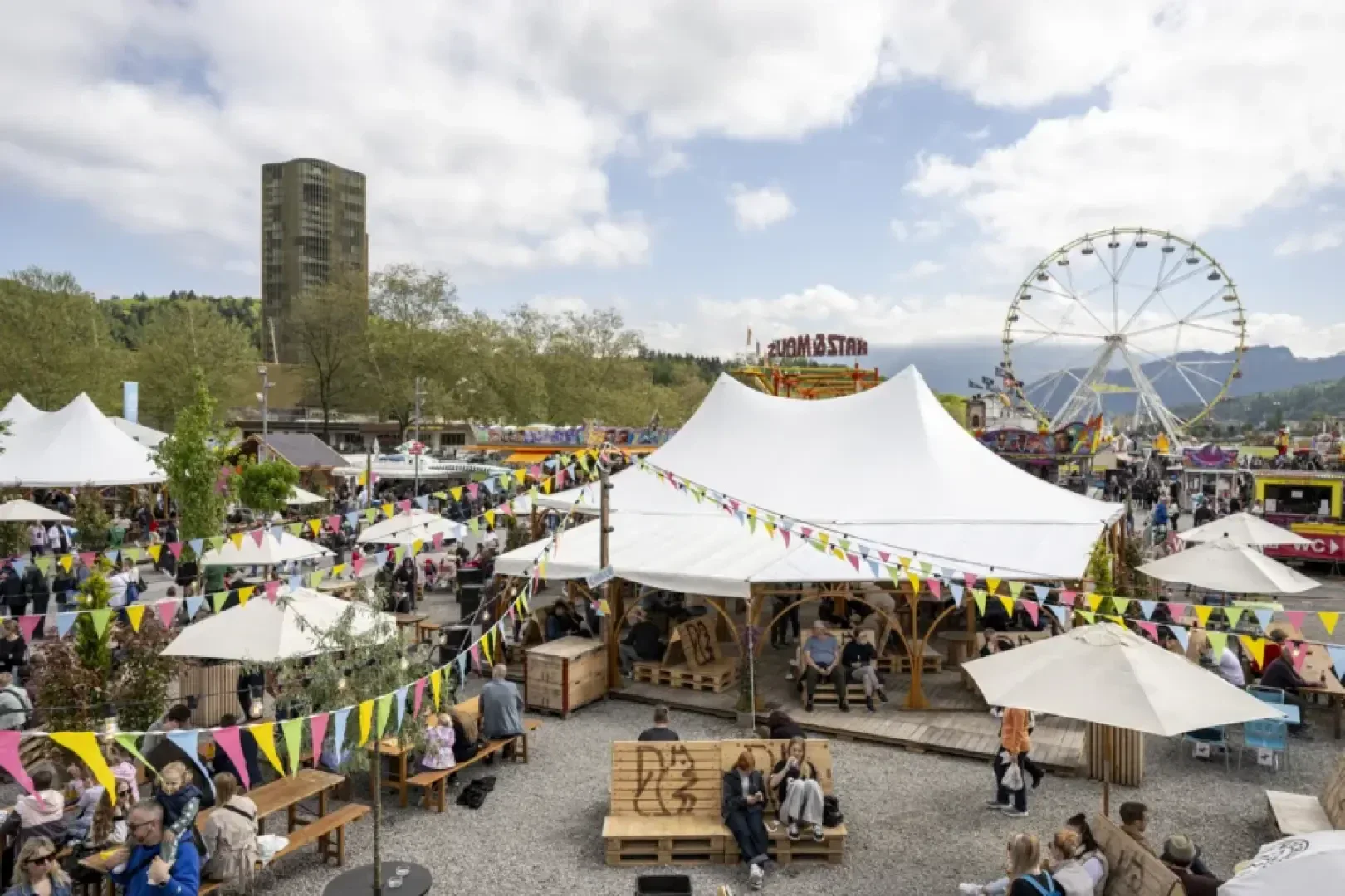 Ein lebhaftes Open-Air-Festival mit weißen Zelten, bunten Wimpeln, Essensständen und herumlaufenden Menschen. Im Hintergrund ist ein Riesenrad zu sehen und links ein hohes Gebäude unter einem teilweise bewölkten Himmel.